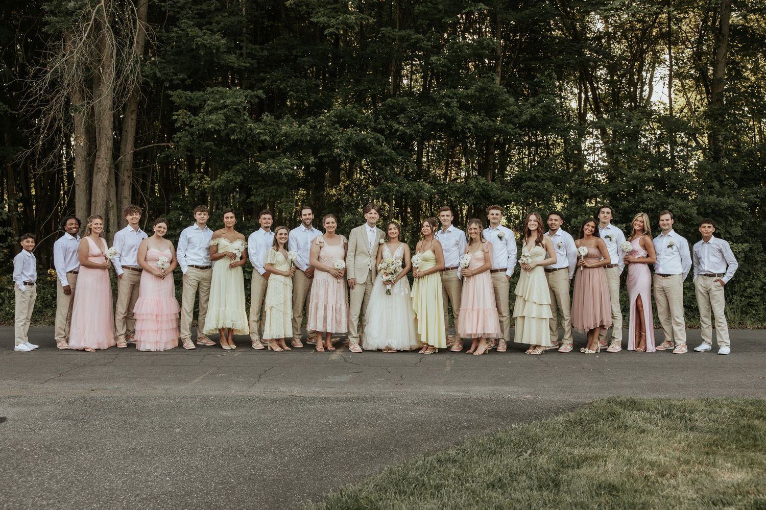 Wedding party members dressed in pink, beige, and white stand together in a wooded outdoor setting for formal photos.