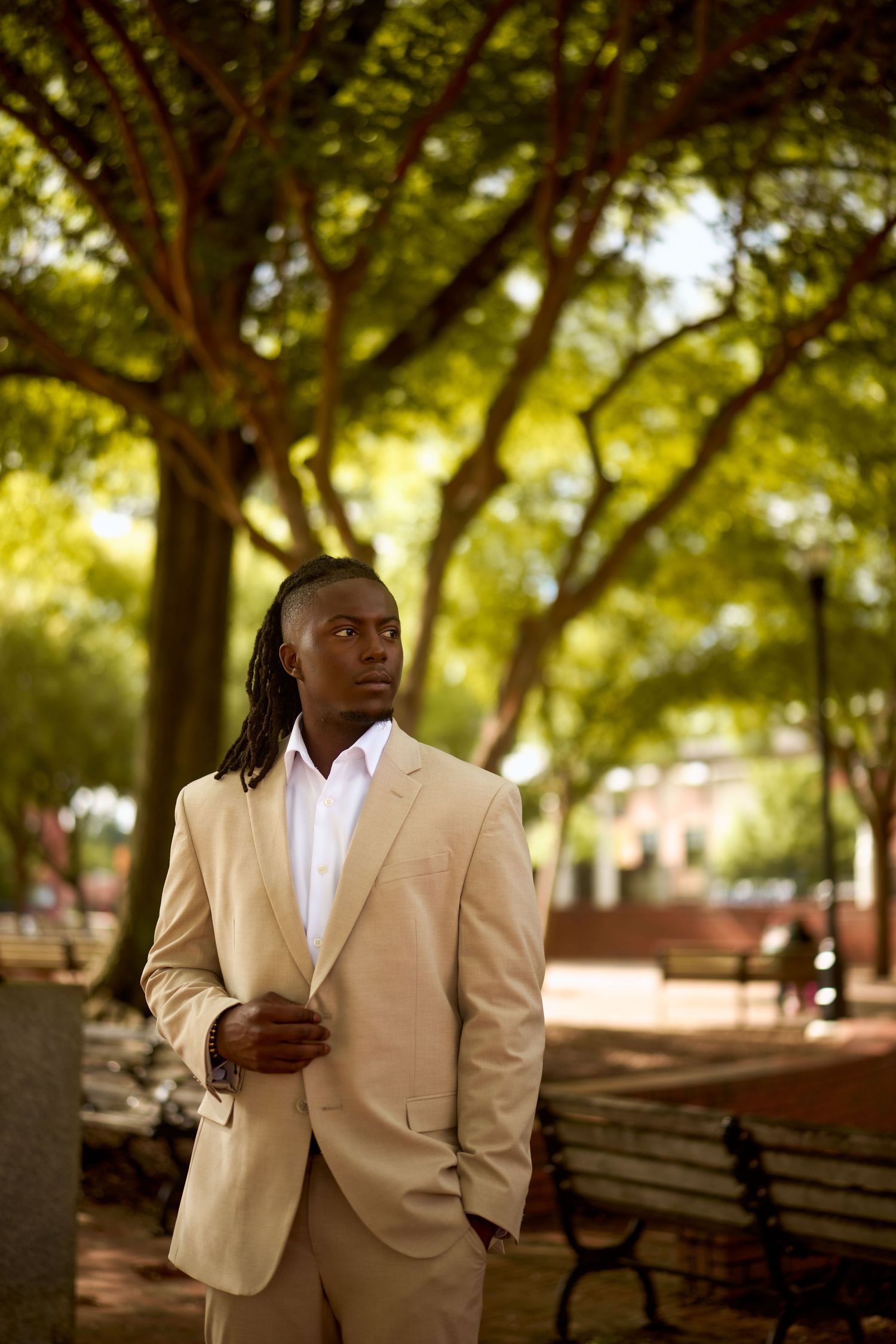 Person in tan suit and white shirt stands thoughtfully in park setting with trees and benches in background.