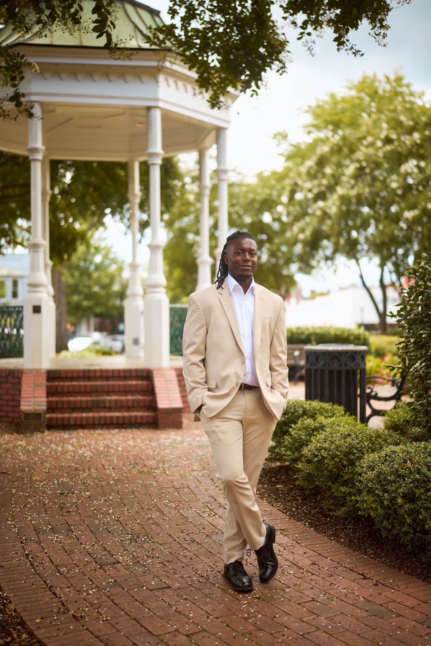 A person in a beige suit stands casually near a white gazebo surrounded by brick walkways and trees.