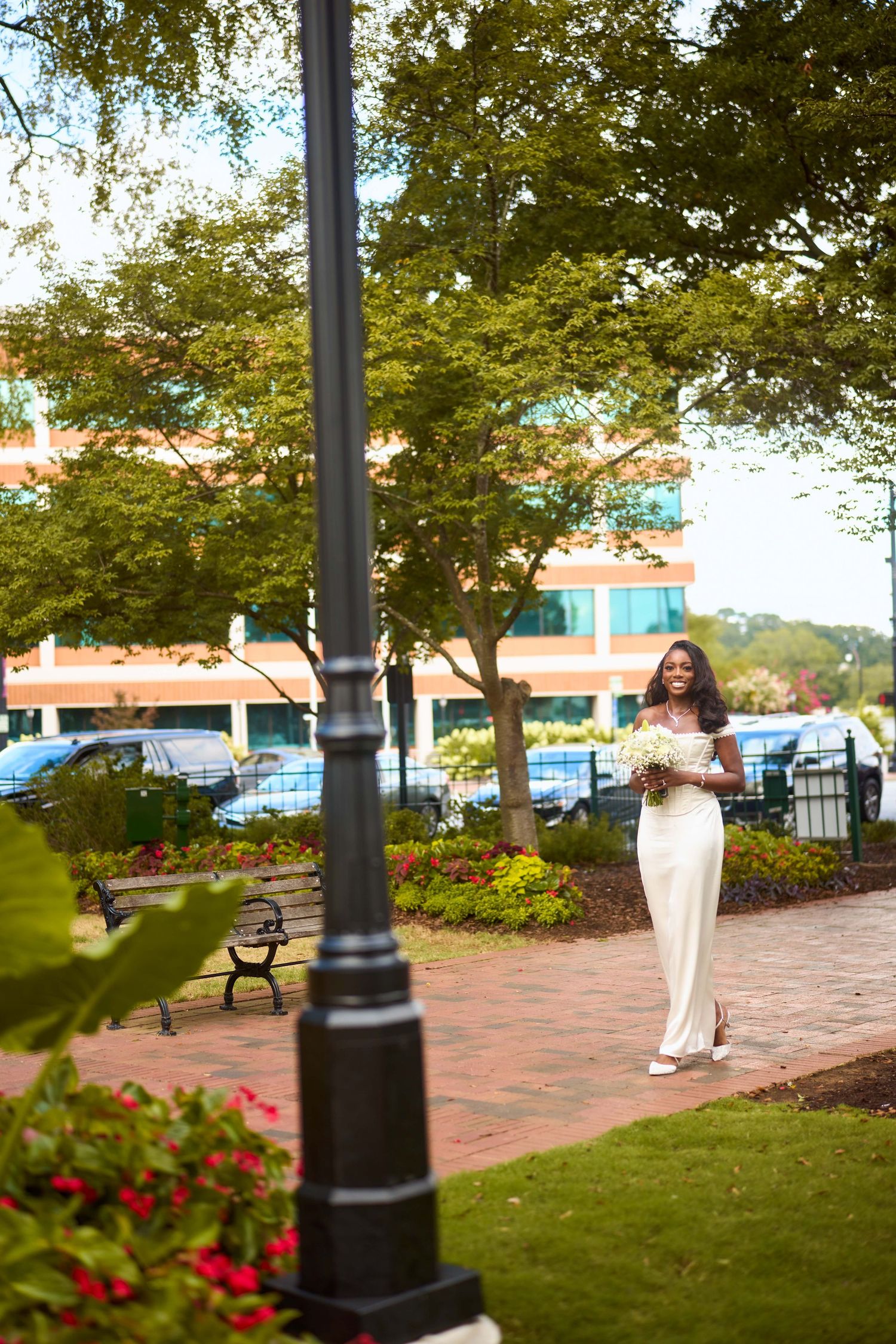 Person in flowing white dress stands among lamp posts and benches on brick path with landscaped greenery.