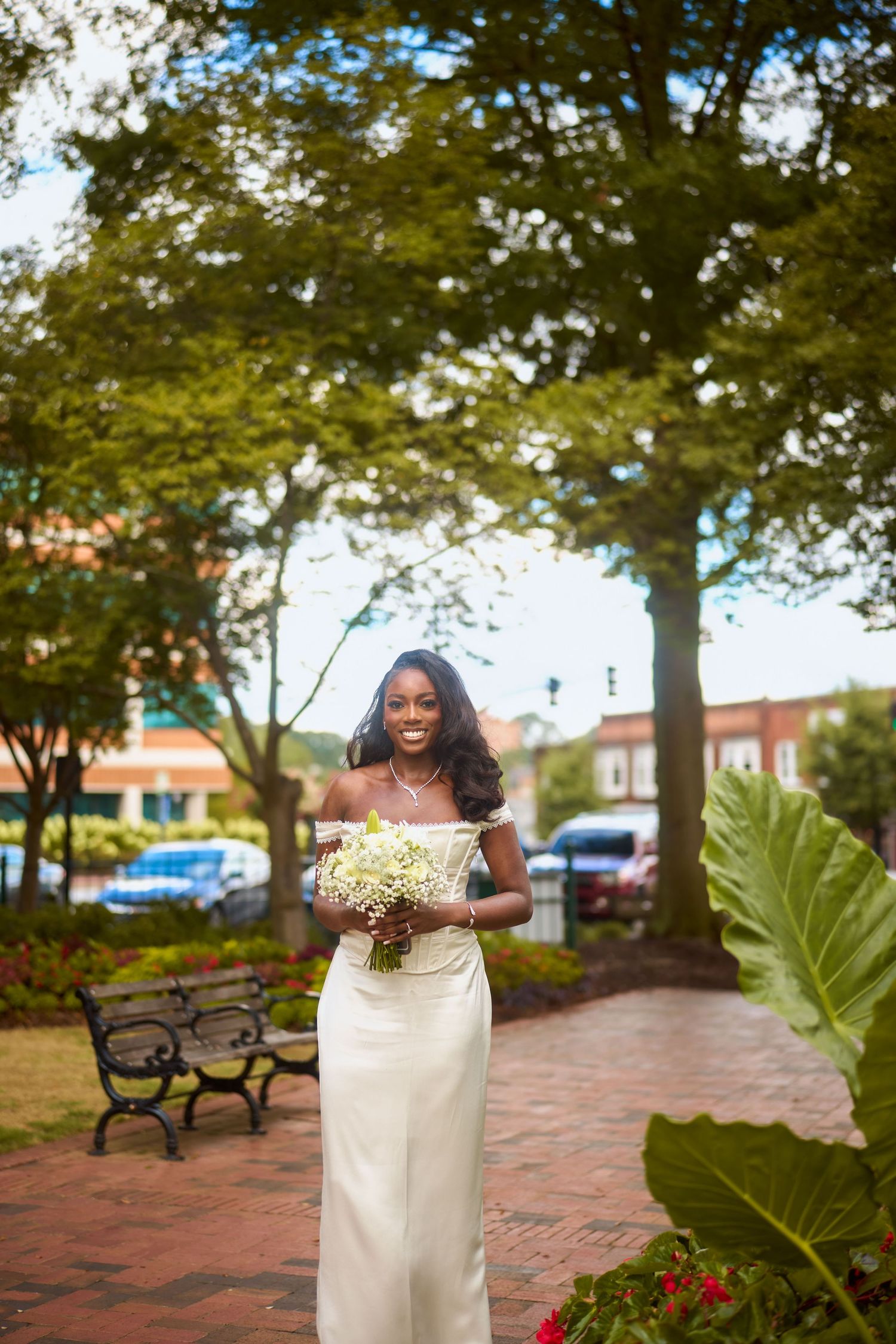 A bride in an elegant off-shoulder white wedding dress holds a bouquet of white flowers in a garden courtyard setting.