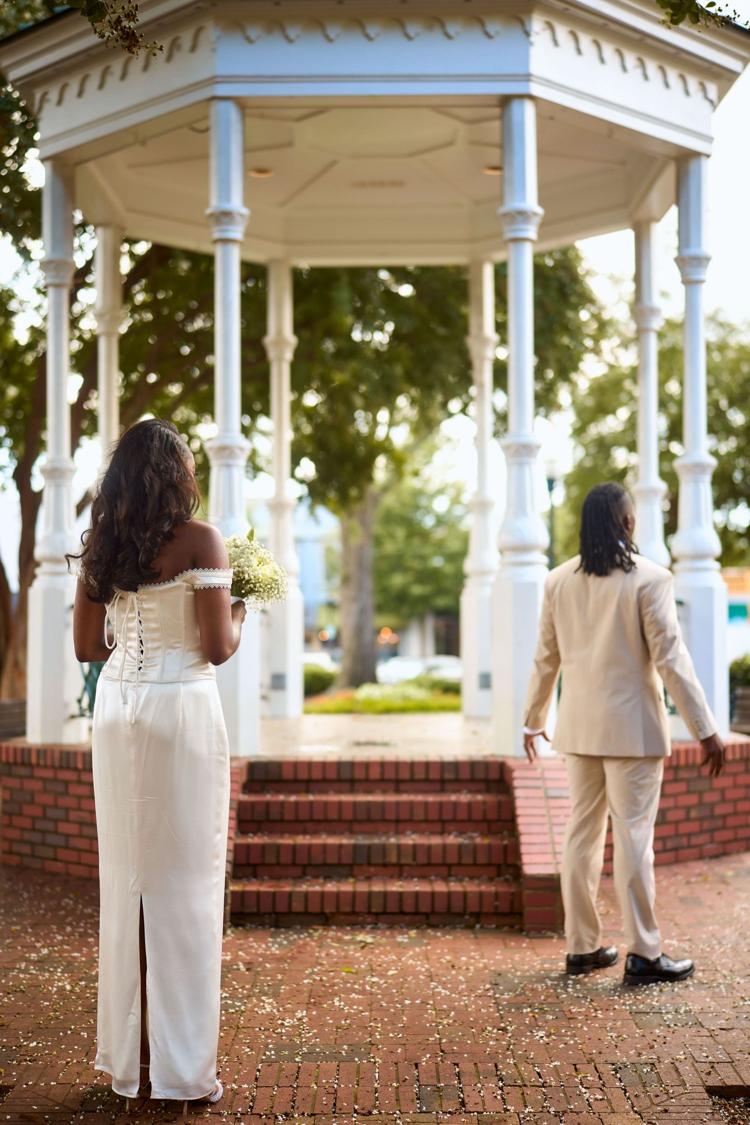 Two people in white outfits stand together in a romantic garden gazebo setting with brick walkways and white columns.