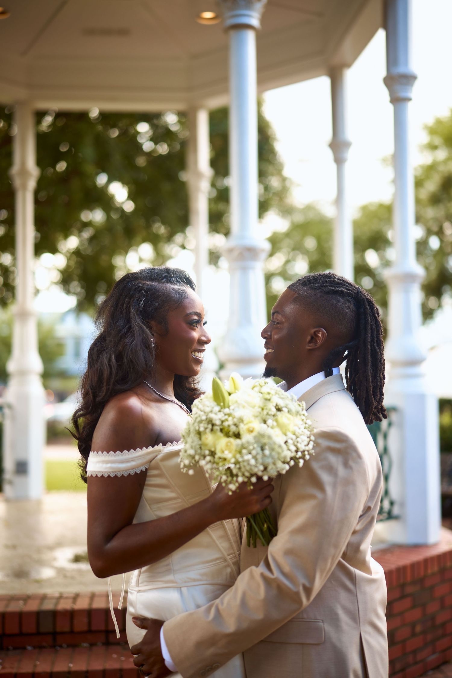 A romantic wedding couple embraces and laughs together under a white columned porch at their outdoor ceremony.