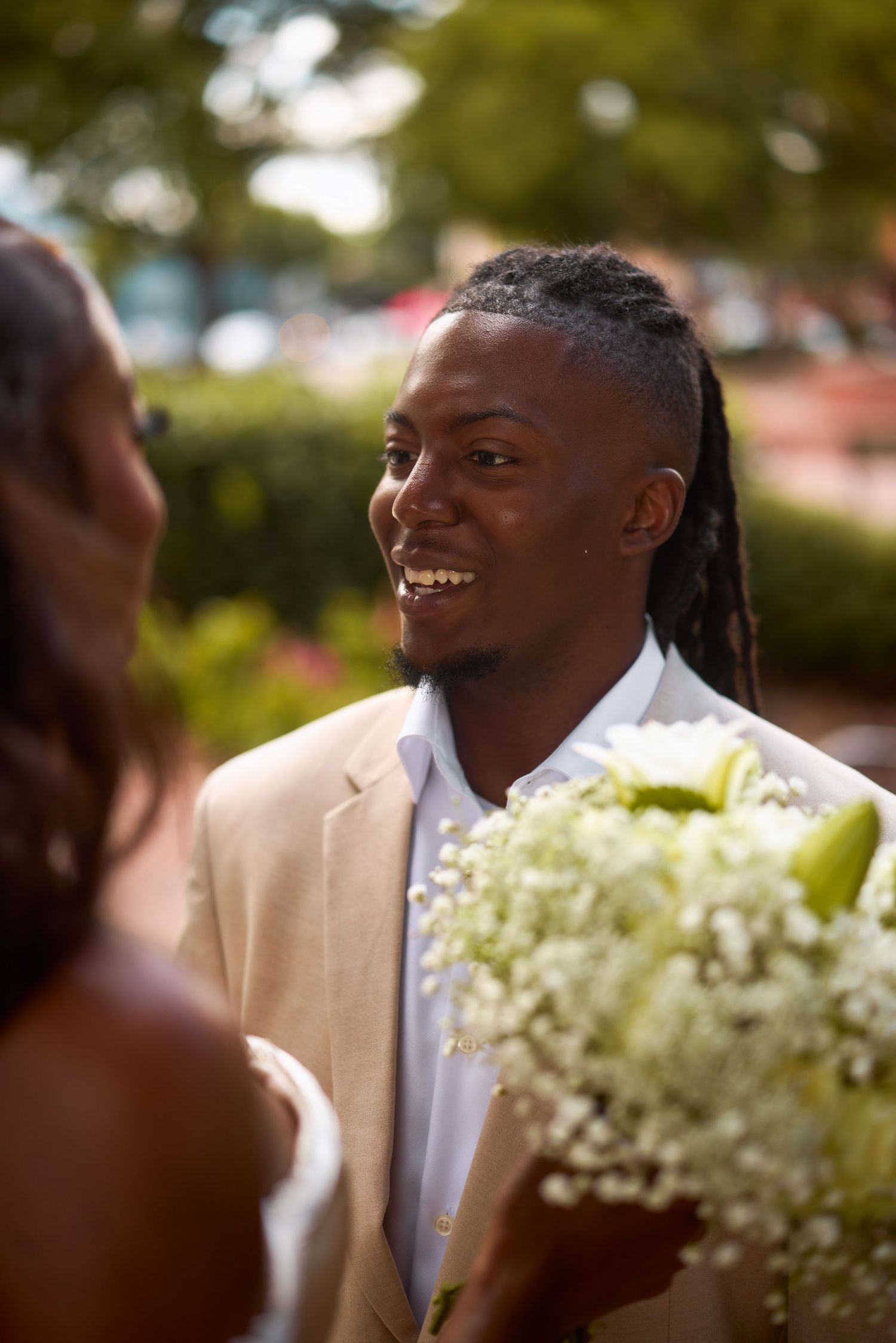 A groom in a beige suit jacket and white shirt smiles warmly during an outdoor wedding ceremony.