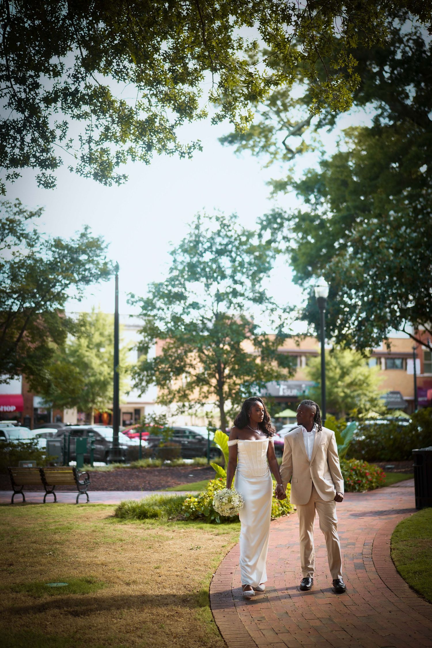 Couple walking hand in hand along a curved brick pathway surrounded by trees and landscaping on a sunny afternoon.