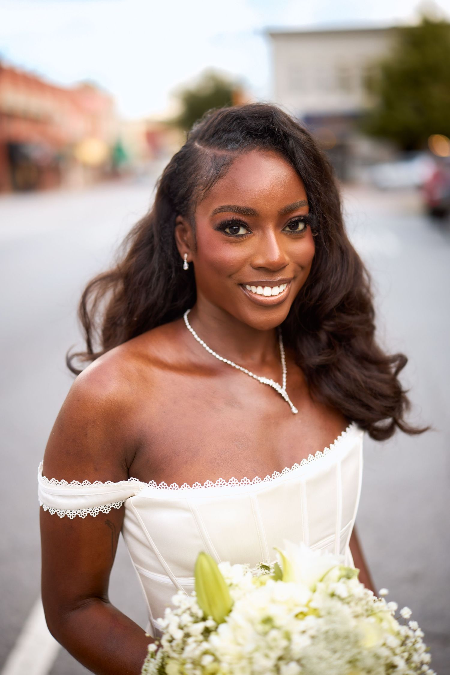 Beautiful bride in off-shoulder white wedding dress holding white and yellow bouquet smiles radiantly outdoors.