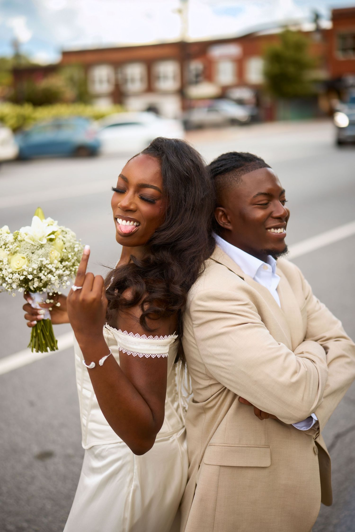 Playful moment between couple in wedding attire laughing and embracing while holding white bouquet outside.