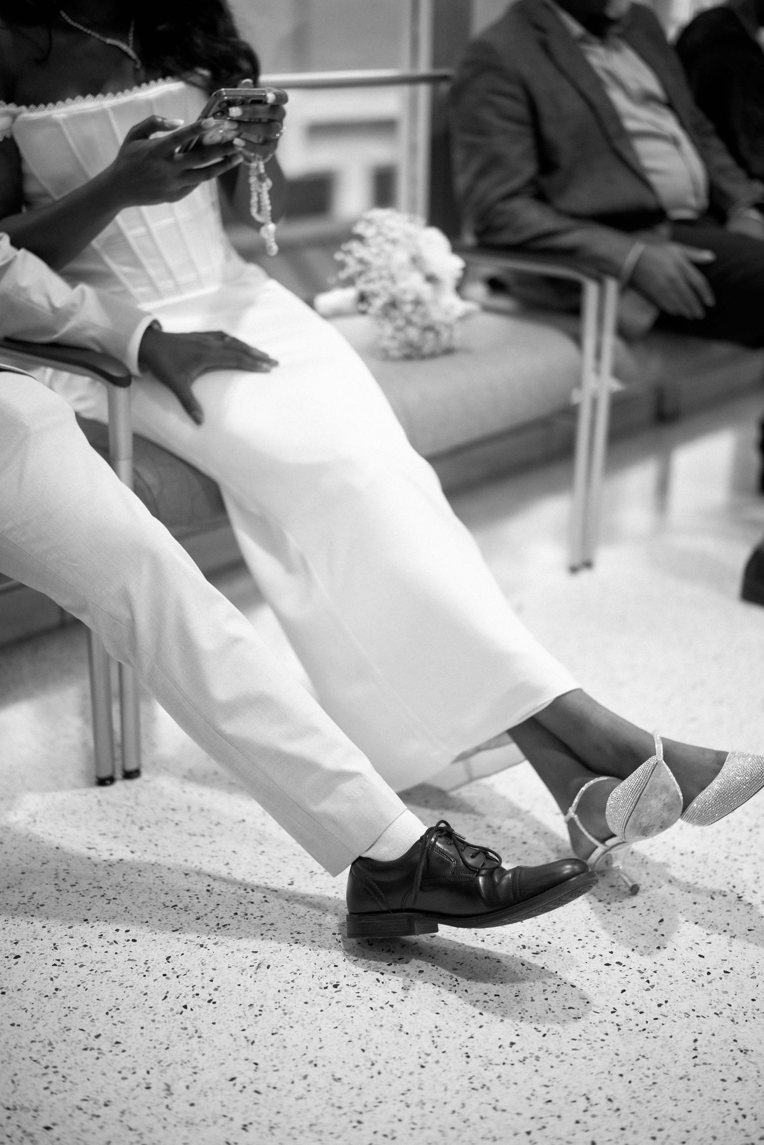 A black and white photo of formal dress shoes and white pants while sitting in a waiting area.