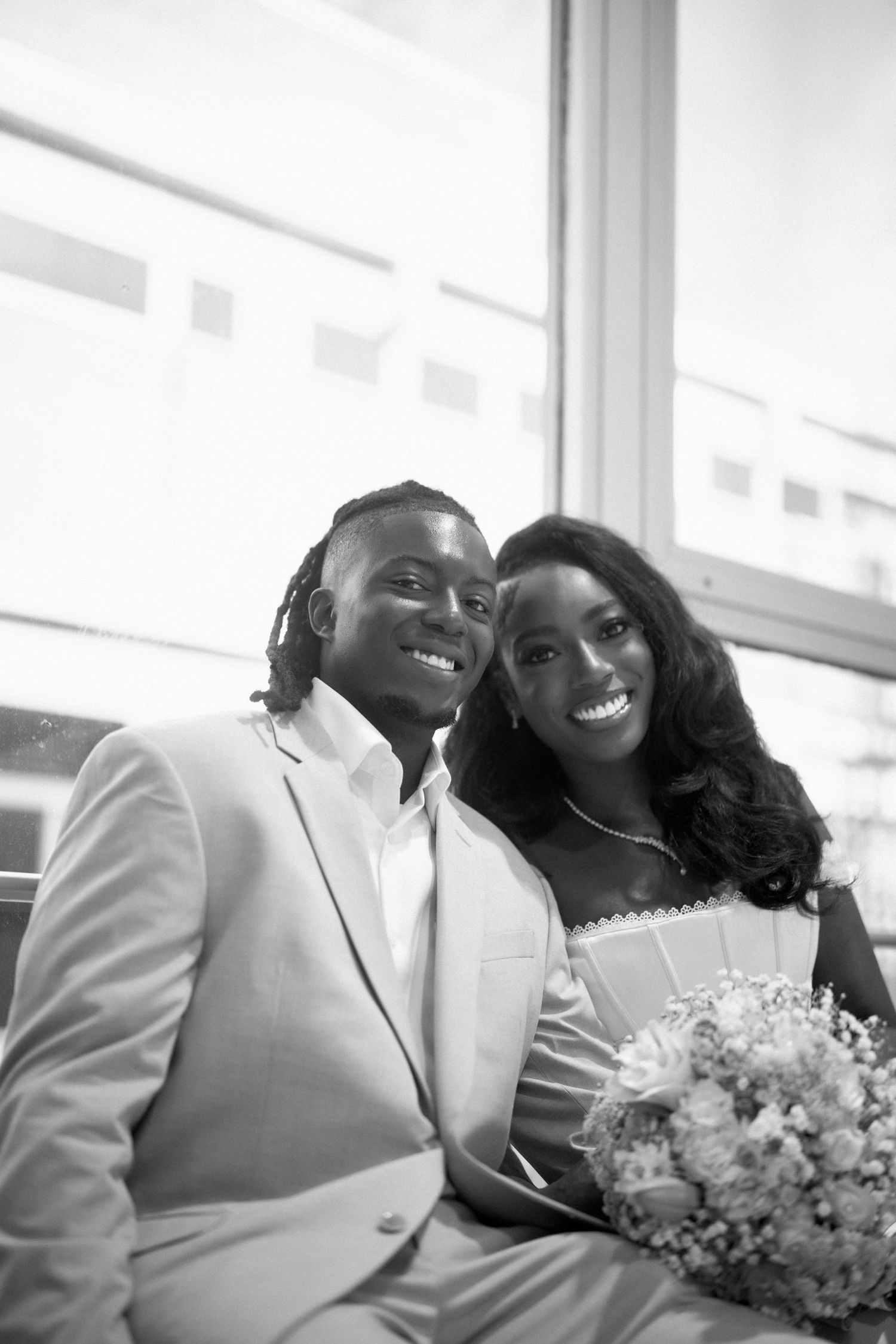 A lovely wedding couple in matching white outfits smiles while sitting together, with the bride holding a delicate bouquet.