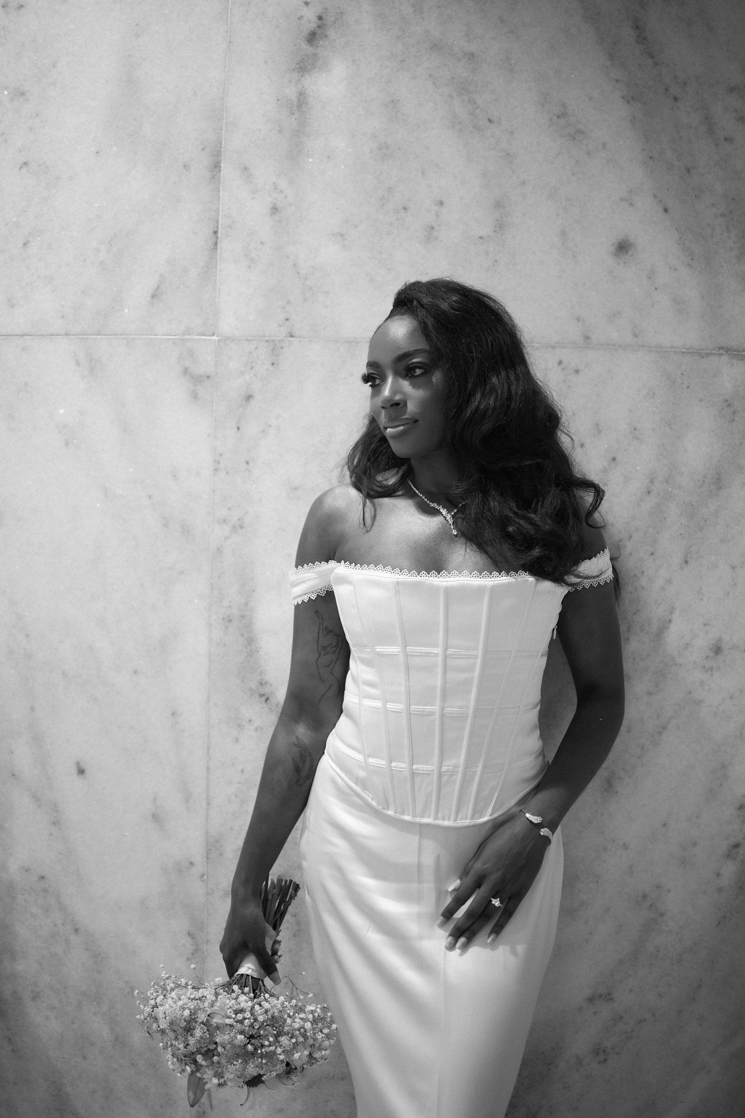 An elegant bride in an off-shoulder corset wedding dress holds a delicate bouquet against a marble wall.