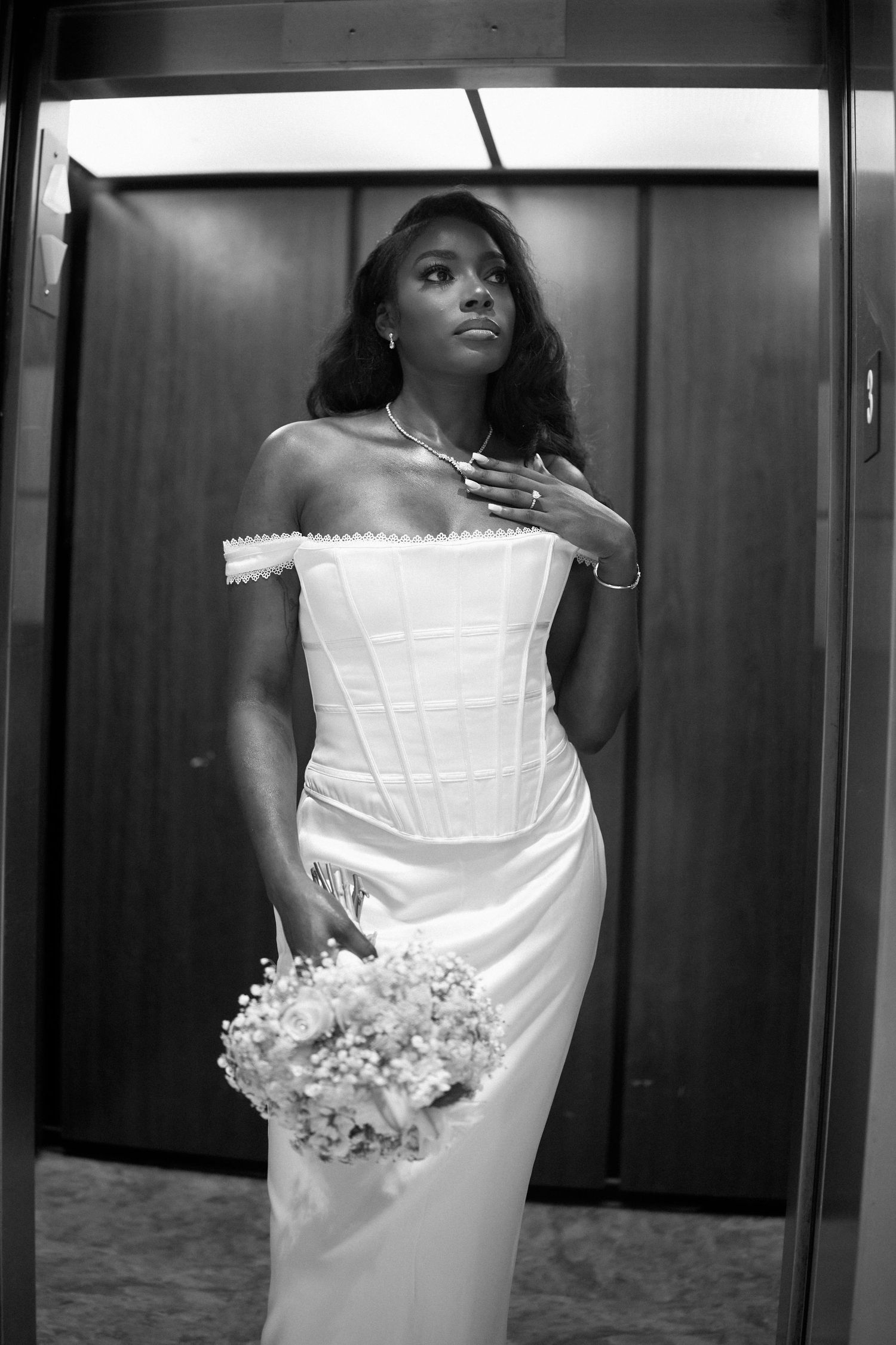 A bride wearing a white corset dress poses with a bouquet in an elevator.