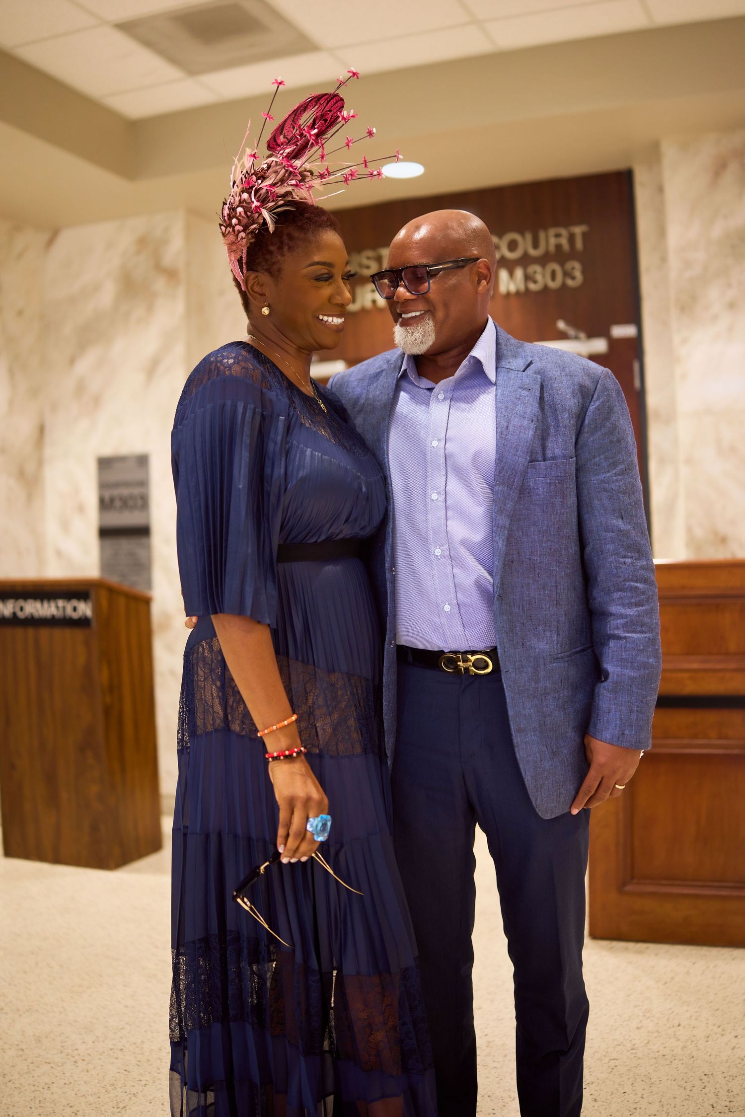 A couple in formalwear shares a tender moment in a courthouse hallway, the woman wearing a navy dress and heeled shoes.