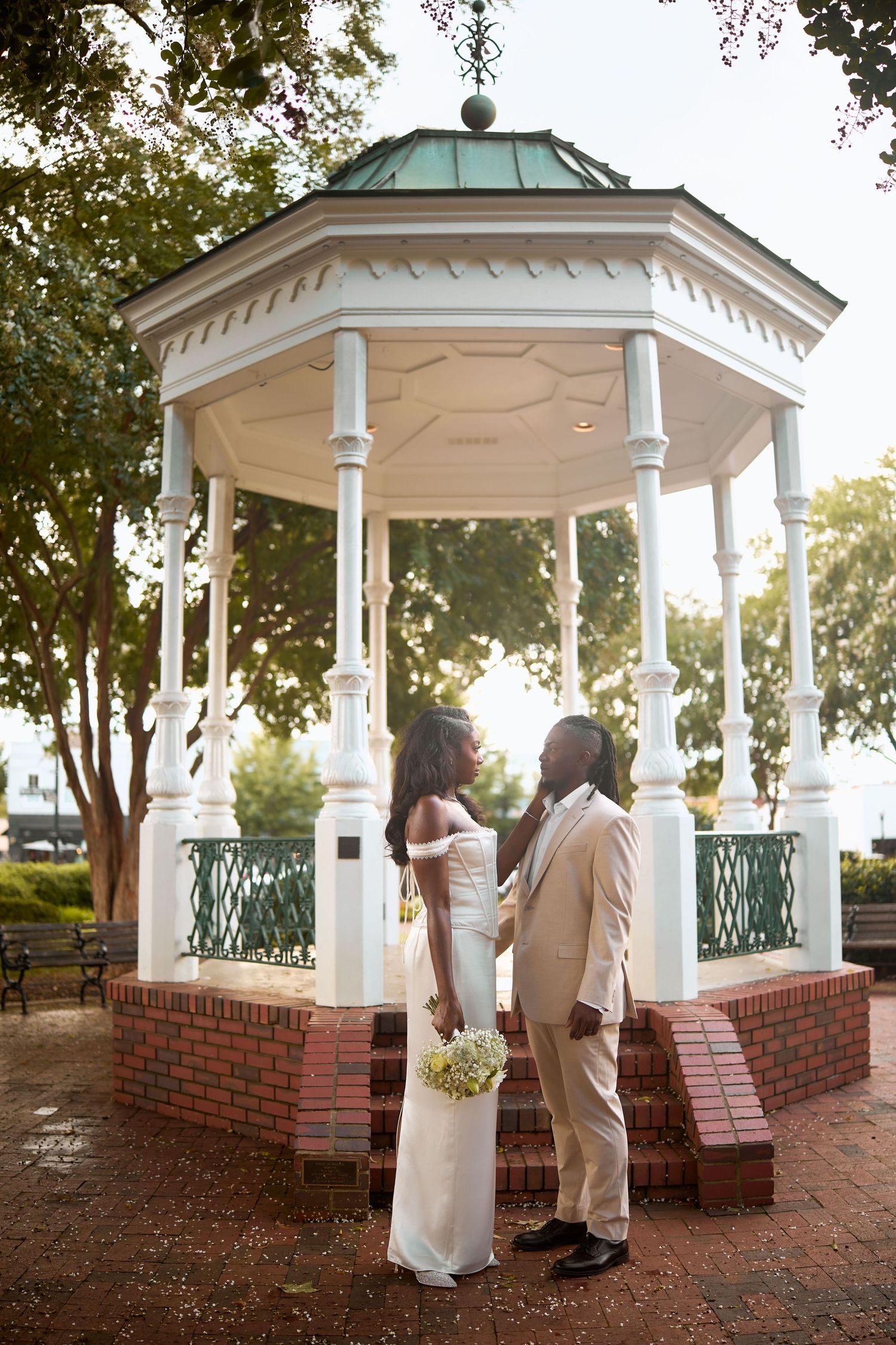 A couple shares an intimate moment under a white gazebo with brick base and green metal railings during a sunset wedding.