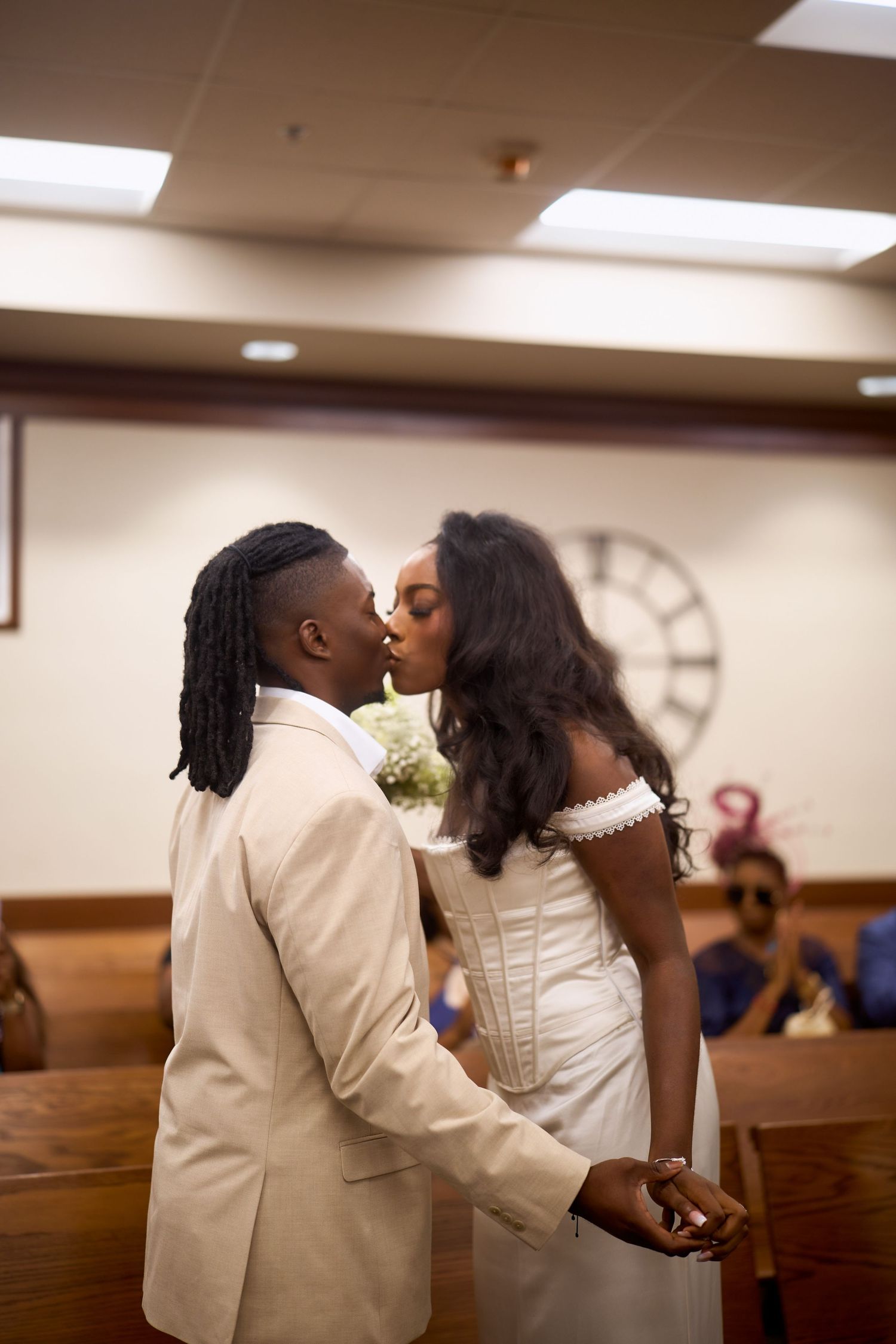 A couple shares their first kiss as newlyweds in a wedding venue with wooden pews and a large clock on the wall.