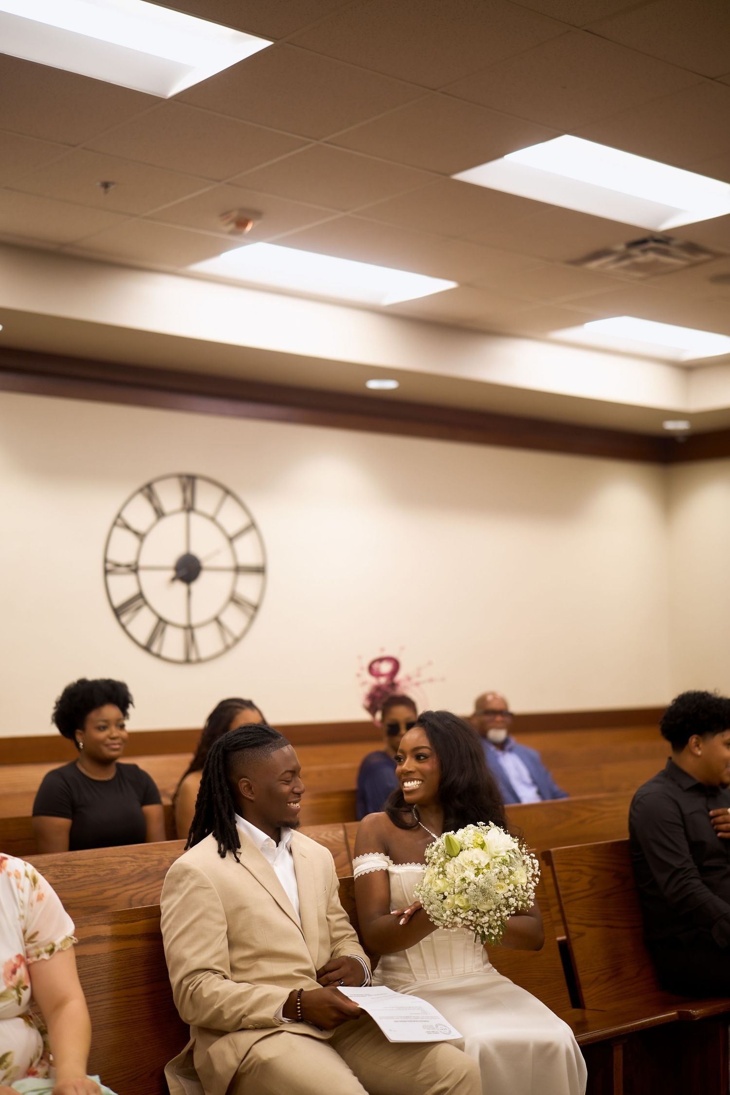 People seated in wooden pews in a room with overhead lighting and a large clock on the wall during a gathering.