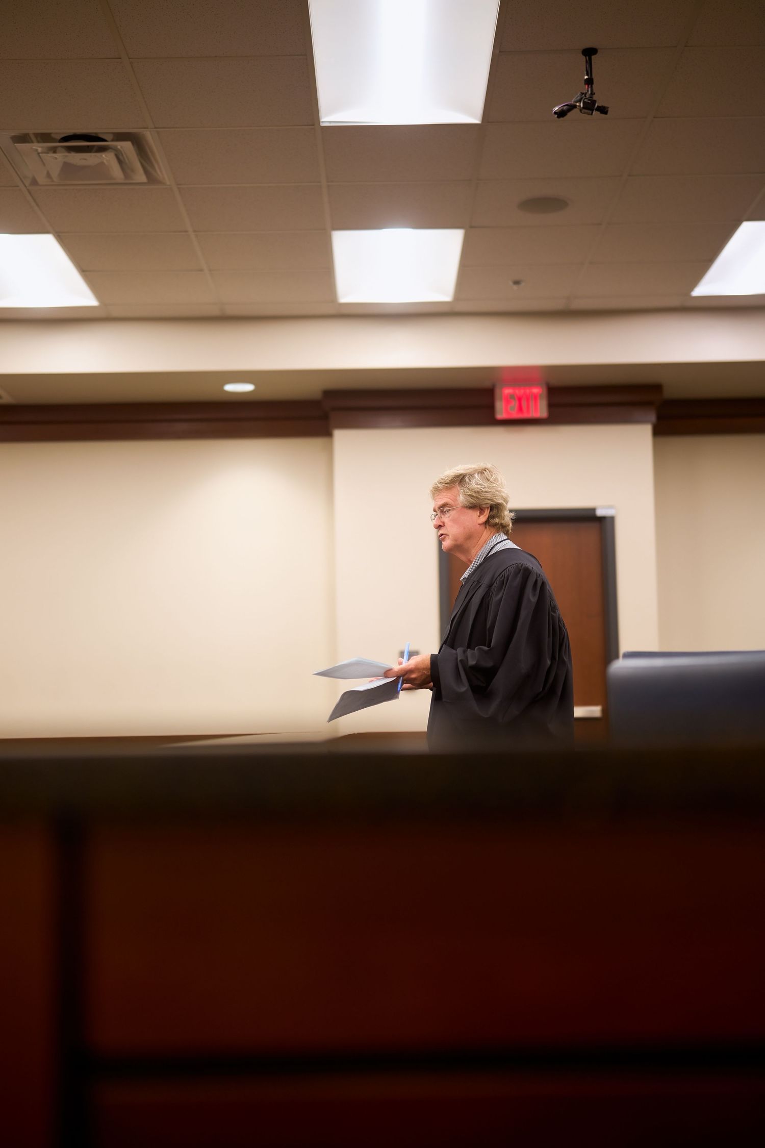 A judge presides over proceedings in a wood-paneled courtroom with bright fluorescent lighting.