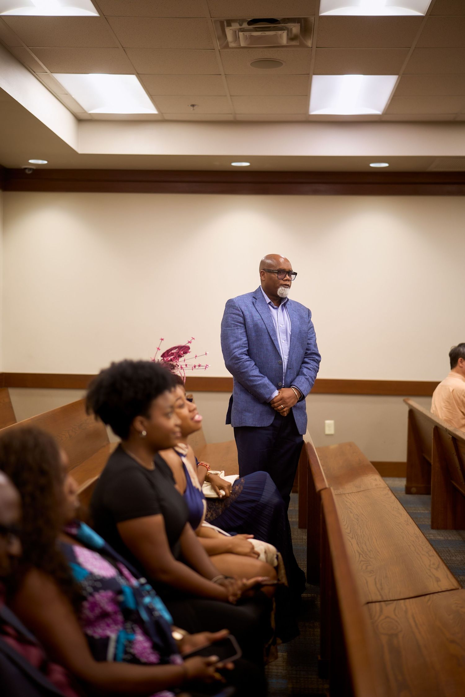 People seated in wooden pews listen attentively during a court session in a brown-toned room.