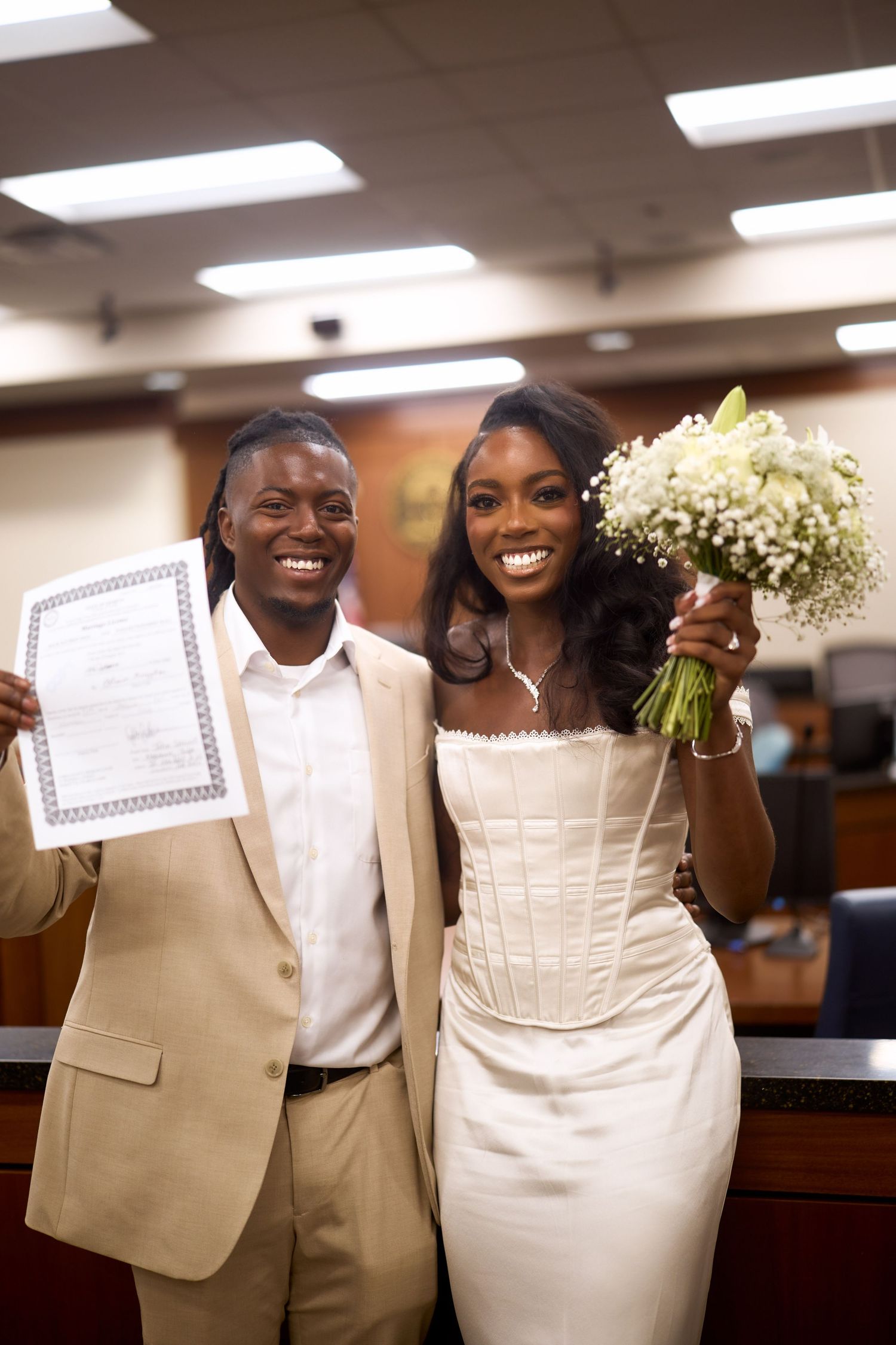 Wedding couple shares a joyful moment while holding paper shopping bags in church reception area.