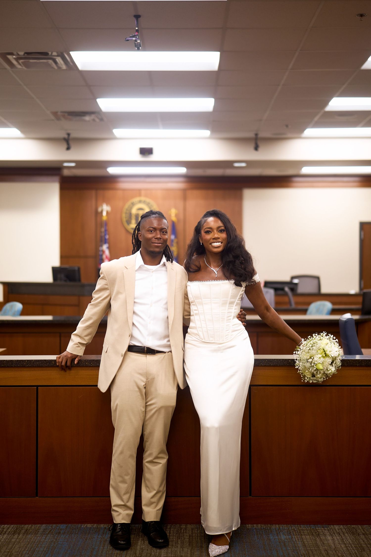 A joyful couple stands together in a courtroom, wearing formal attire of tan and white for their civil ceremony.