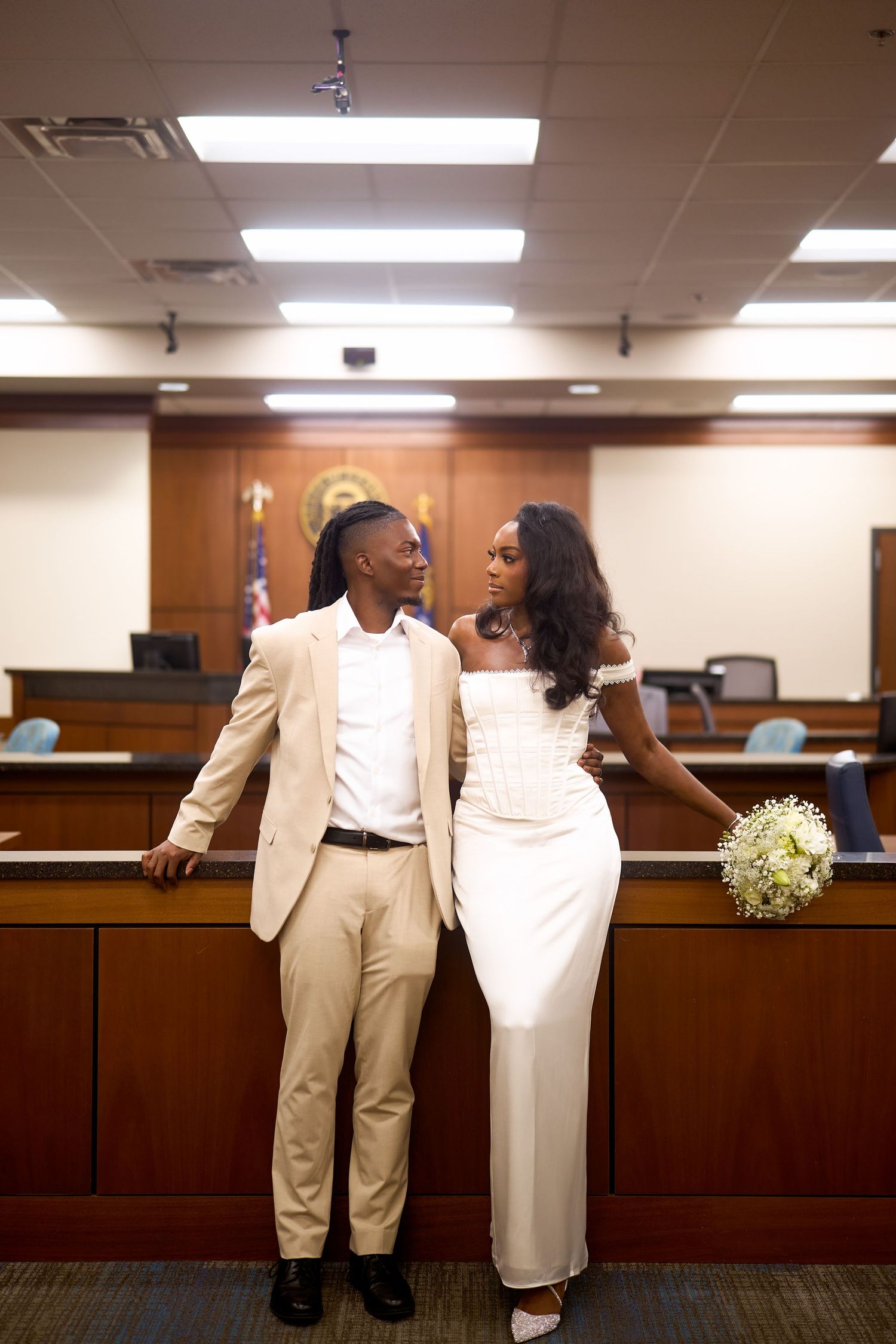 A couple dressed in matching white and tan formal attire smiles at each other in a courthouse setting.