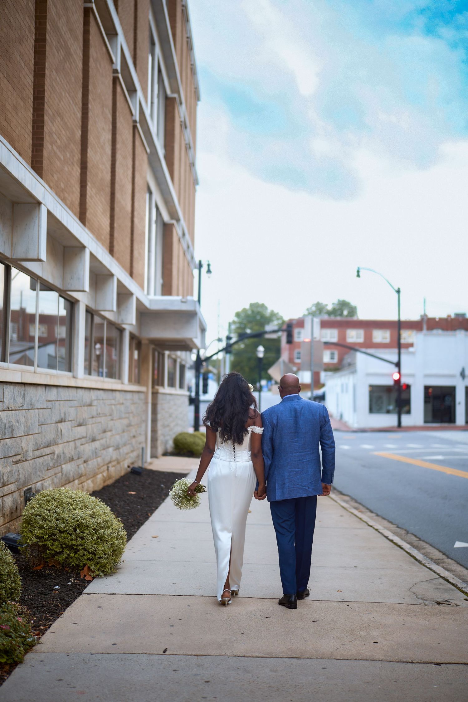 Two figures walk together down an urban sidewalk beside a brick apartment building on a sunny day.