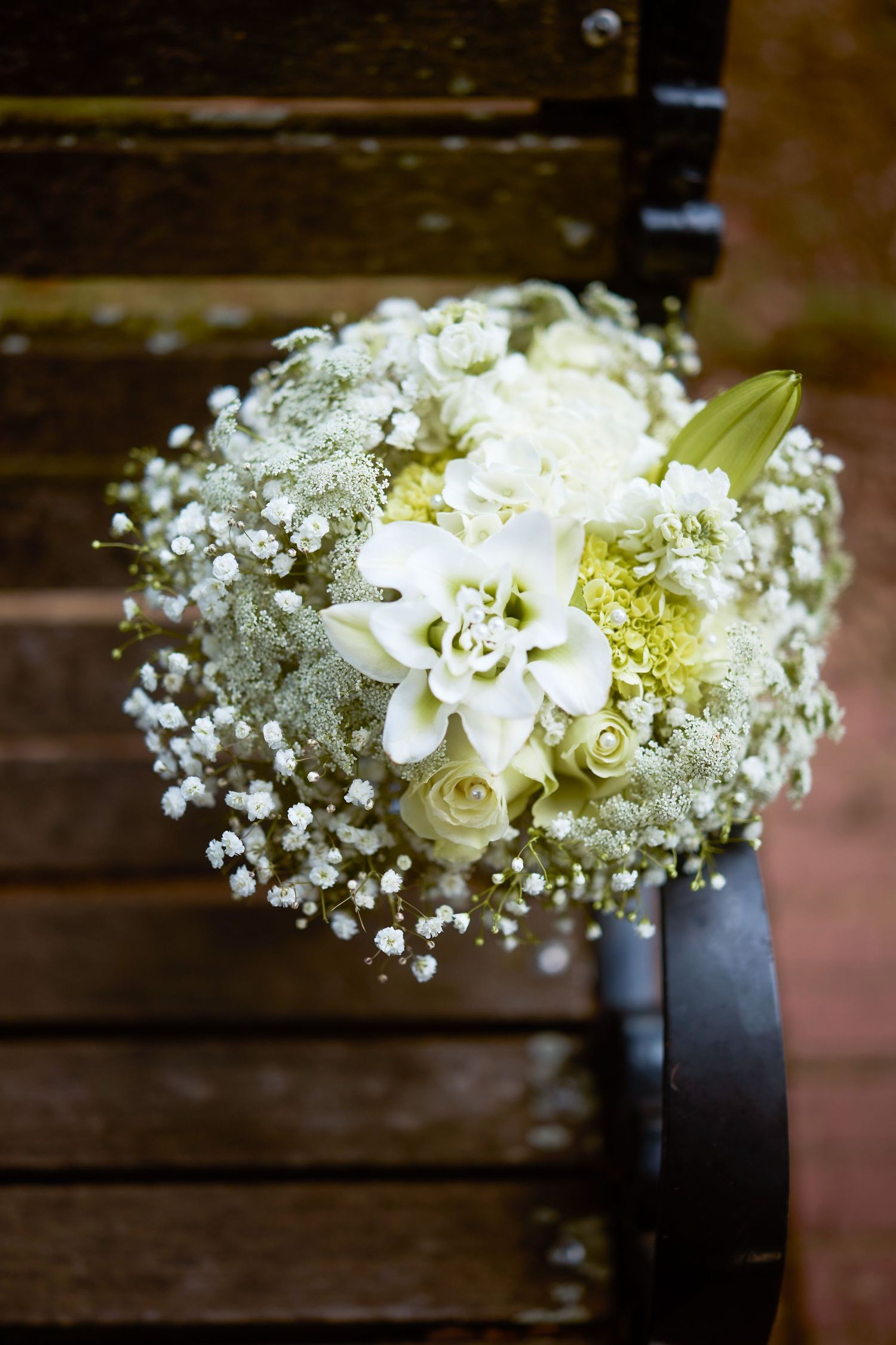 Delicate white wedding bouquet featuring roses and baby's breath arranged with black ribbon on wooden surface.