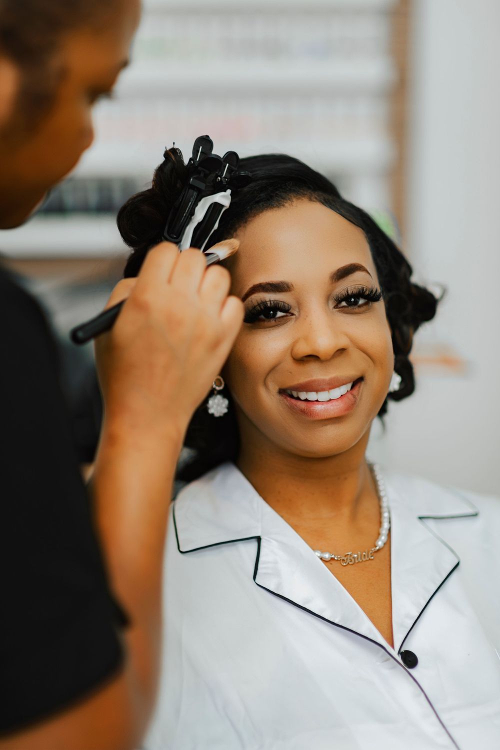 Makeup artist applies cosmetics to a smiling client wearing a white collared outfit and sparkly necklace.