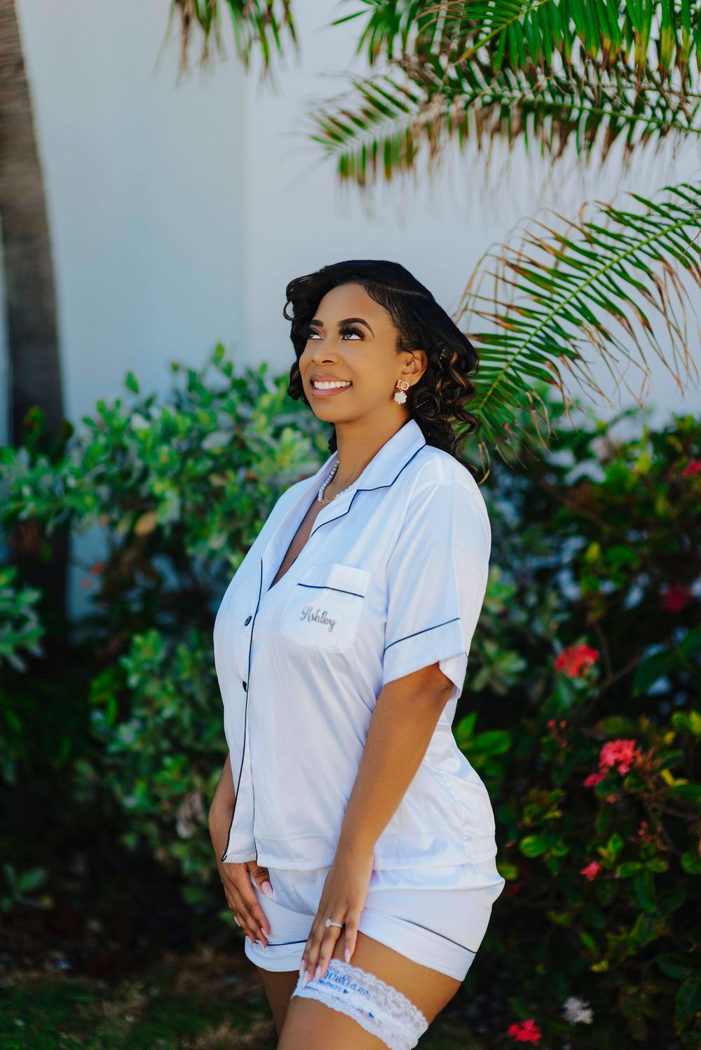 Person in white uniform poses outdoors against tropical garden backdrop with palm fronds and red flowers.