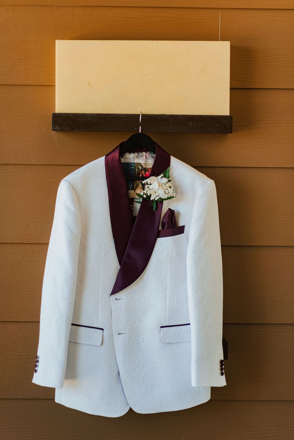 White formal blazer with burgundy lapel and floral boutonniere displayed against wooden paneled wall.