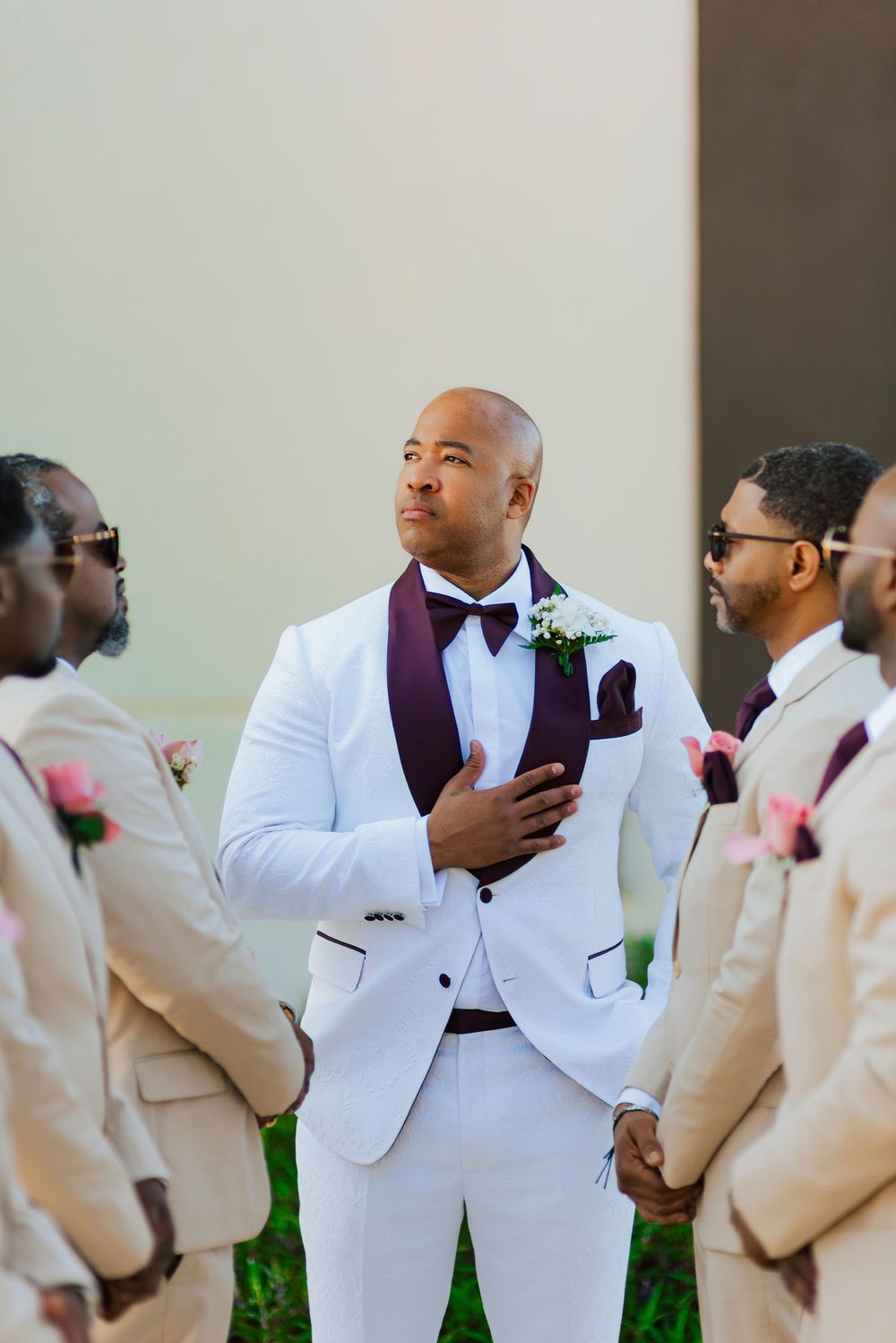 Groomsmen in crisp white tuxedo jackets and burgundy bowties gather together for wedding party photos outdoors.