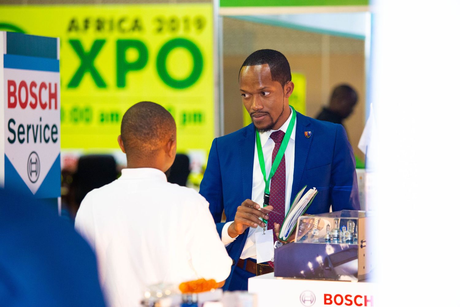Business professionals in blue suits examine automotive technology displays at an industry expo event.