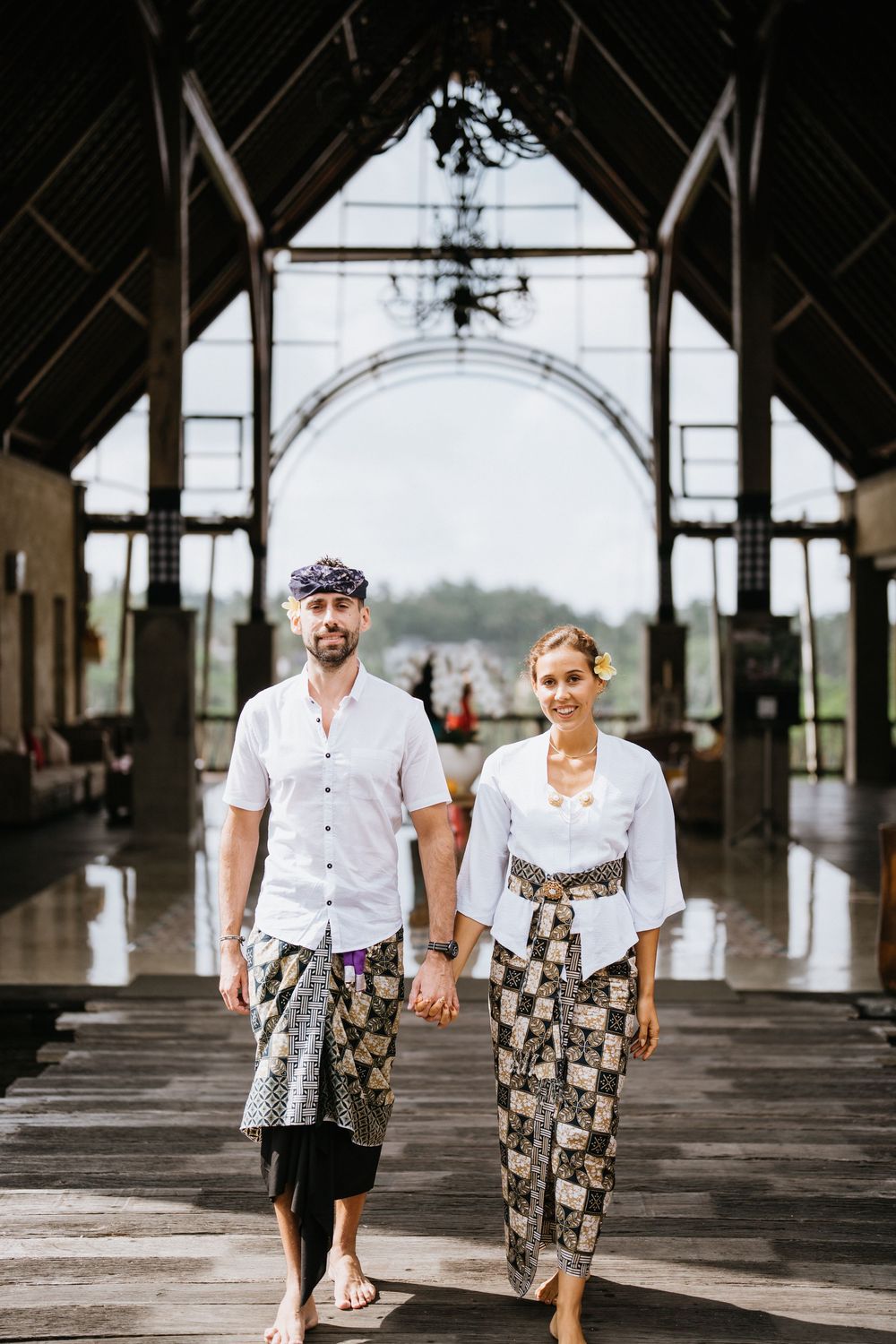 Two people in coordinated white tops and batik bottoms walk through a covered wooden pavilion.