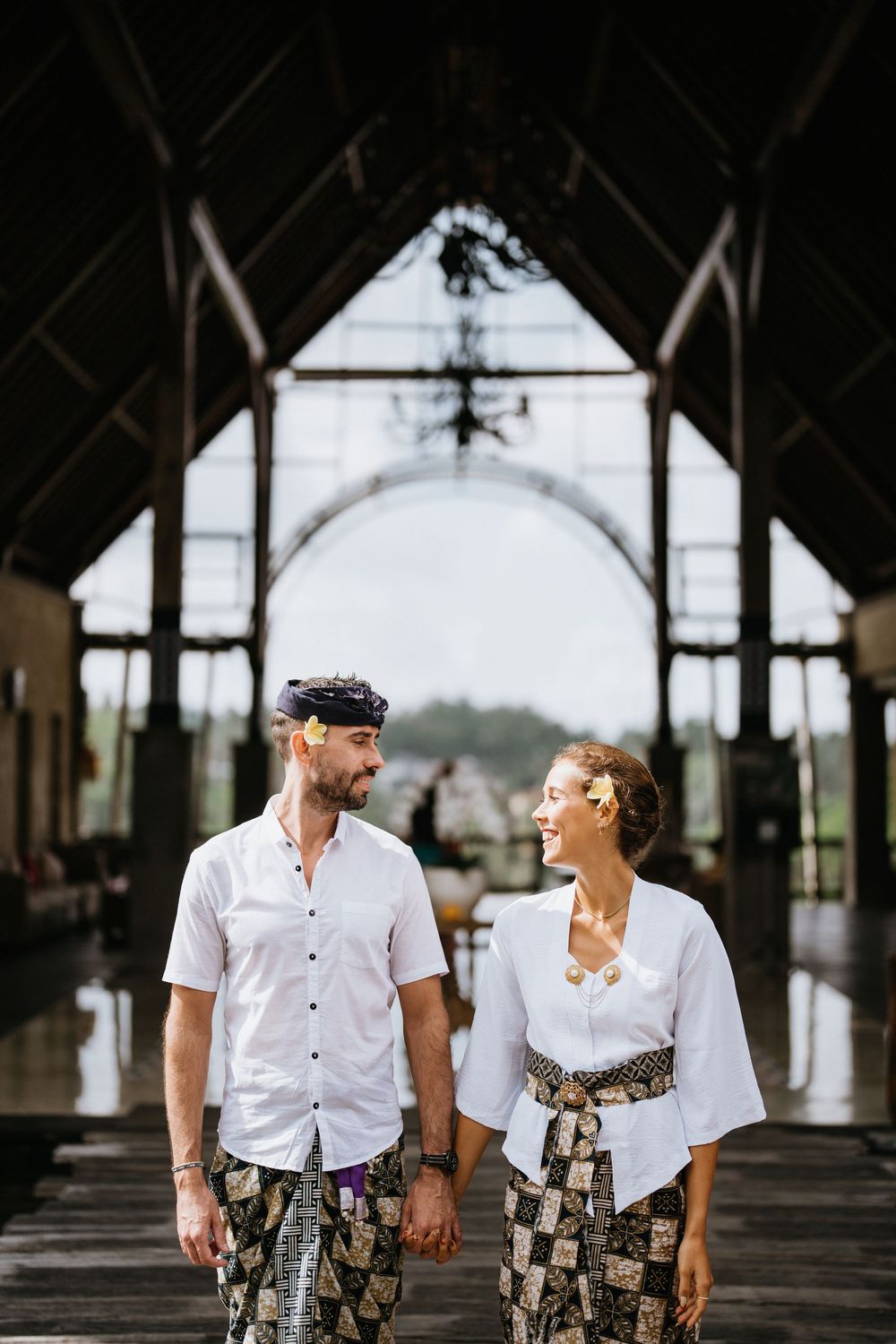A couple walks along a covered wooden walkway wearing matching white shirts and batik patterns.