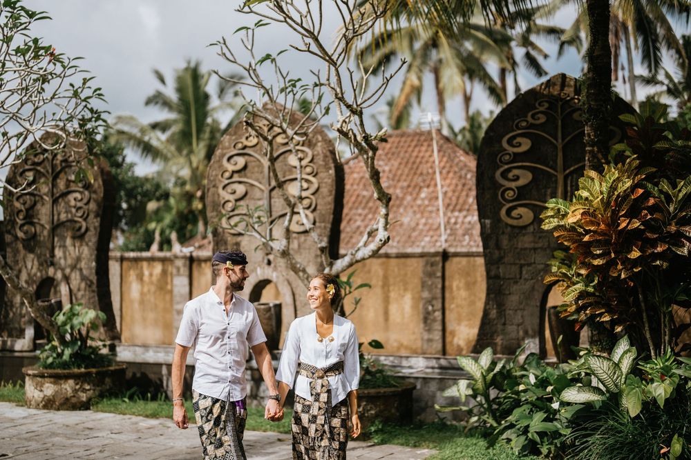 A couple strolls through a tropical garden with traditional Balinese architecture in the background.