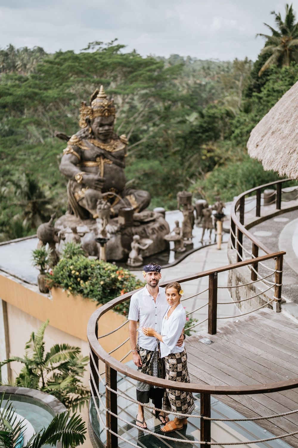 A couple stands on a wooden deck overlooking a tropical valley with stone statues.