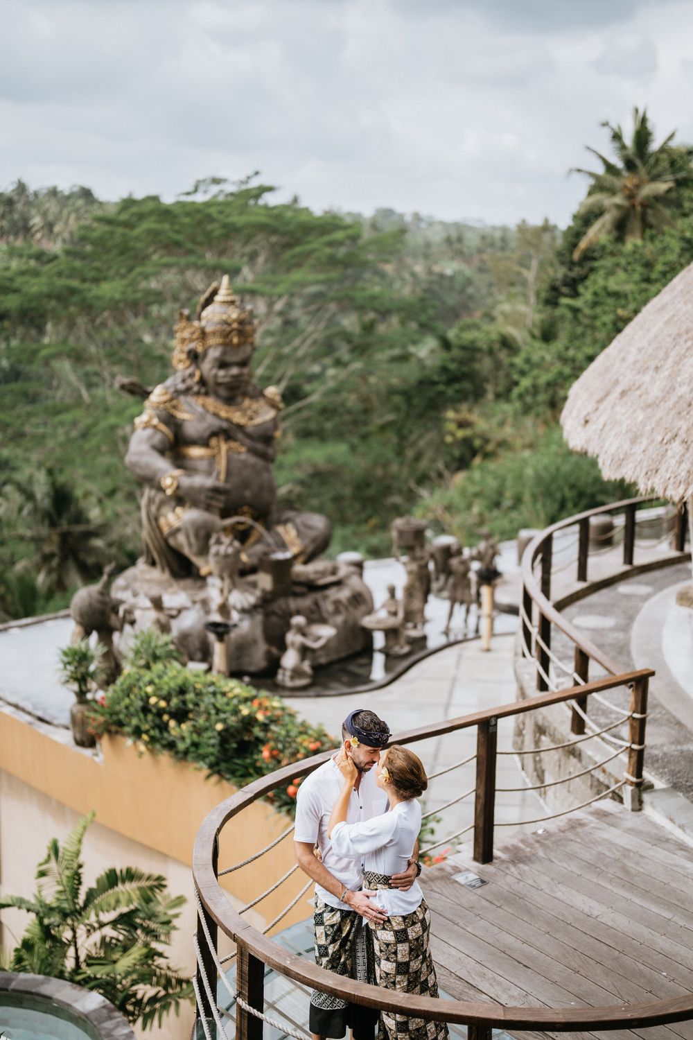Two people embrace on a wooden deck with Balinese statues and tropical landscape in background.