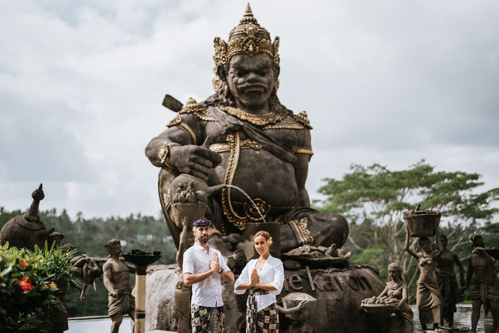 Two people stand near large ornate Balinese stone statues on a tropical hillside.