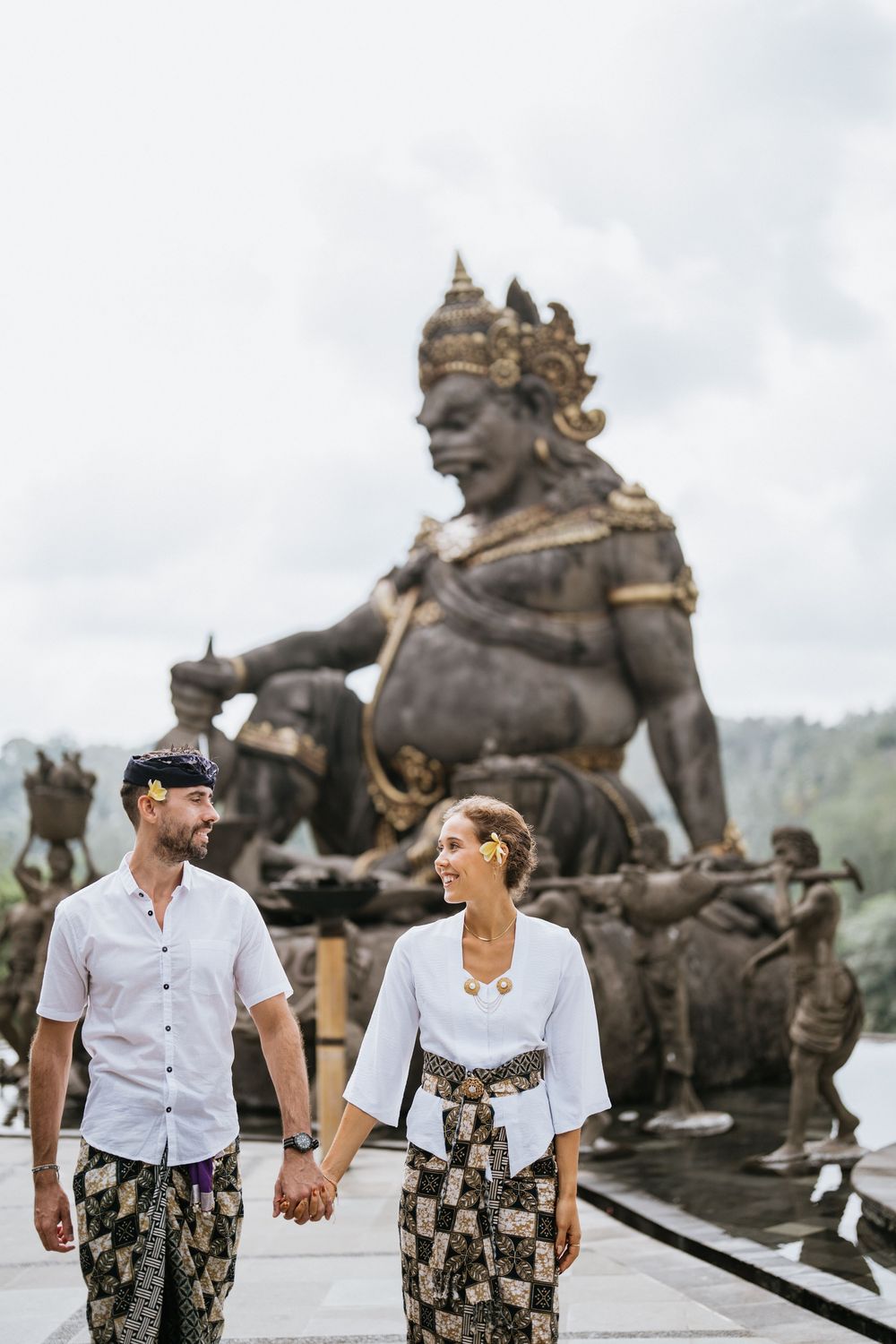 Couple in matching white shirts and floral pants pose by traditional Balinese statues at a luxury resort.