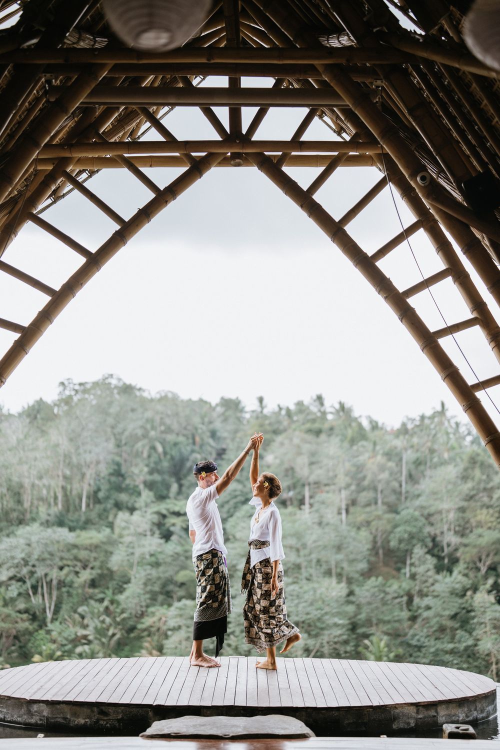 Couple dancing under bamboo roof structure with tropical forest backdrop.