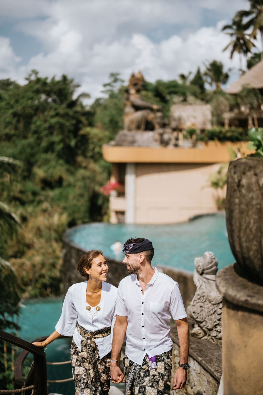 Couple enjoying scenic views from resort terrace with stone statues and tropical landscape.