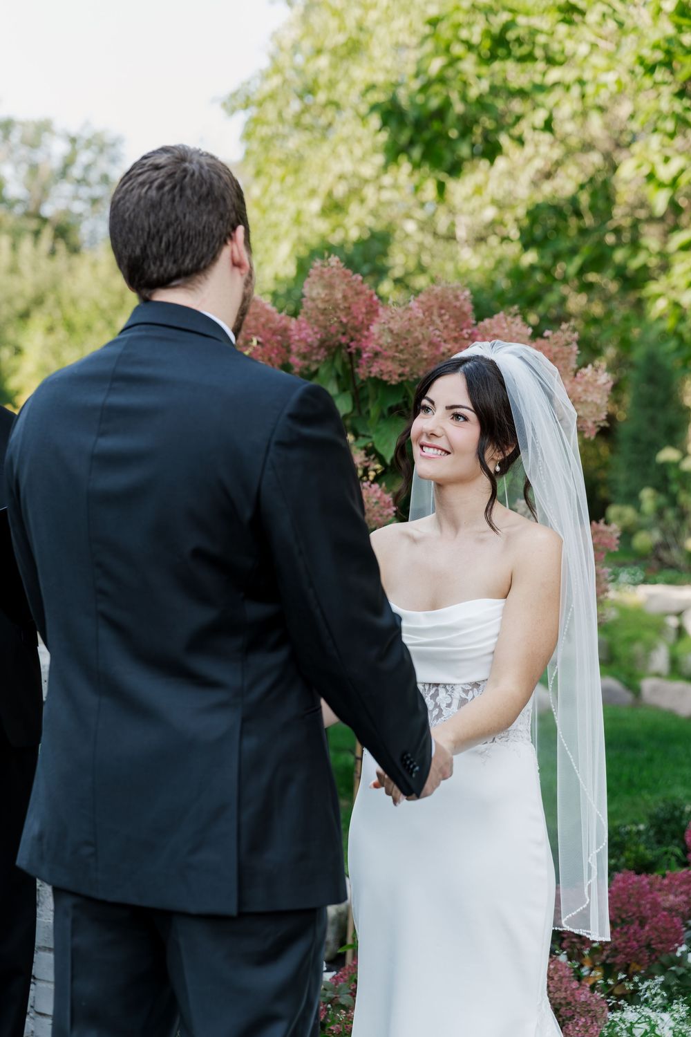 Bride smiles up at her groom during their garden ceremony surrounded by greenery at The Wren House in St. Jacobs, Ontario.