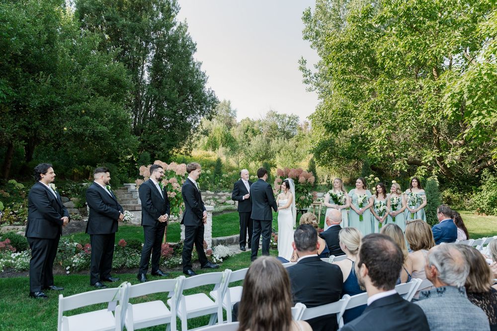 Bride and groom and wedding party stand in front of guests in the gardens surrounded by trees at The Wren House in St. Jacobs, Ontario.