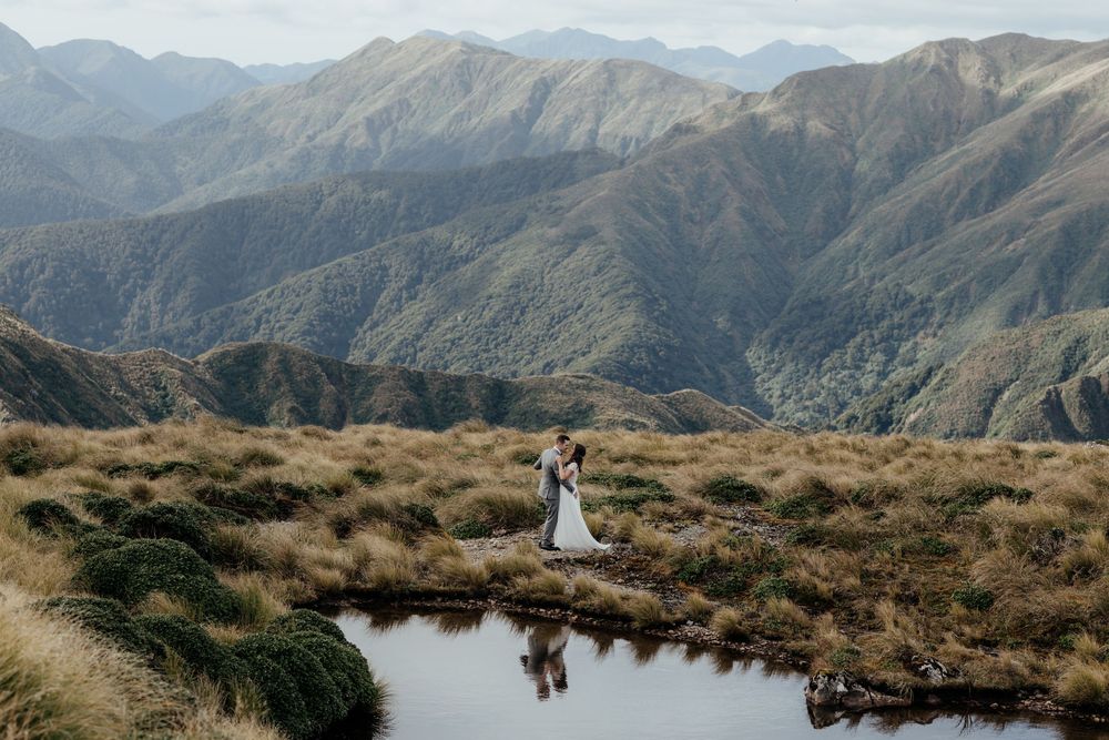 A bride and groom embrace by an alpine lake in the rugged mountain landscape of the Tararua Ranges in Wairarapa New Zealand by August & Vine