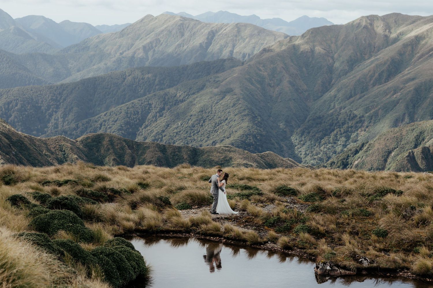 A bride and groom embrace by an alpine lake in the rugged mountain landscape of the Tararua Ranges in Wairarapa New Zealand by August & Vine