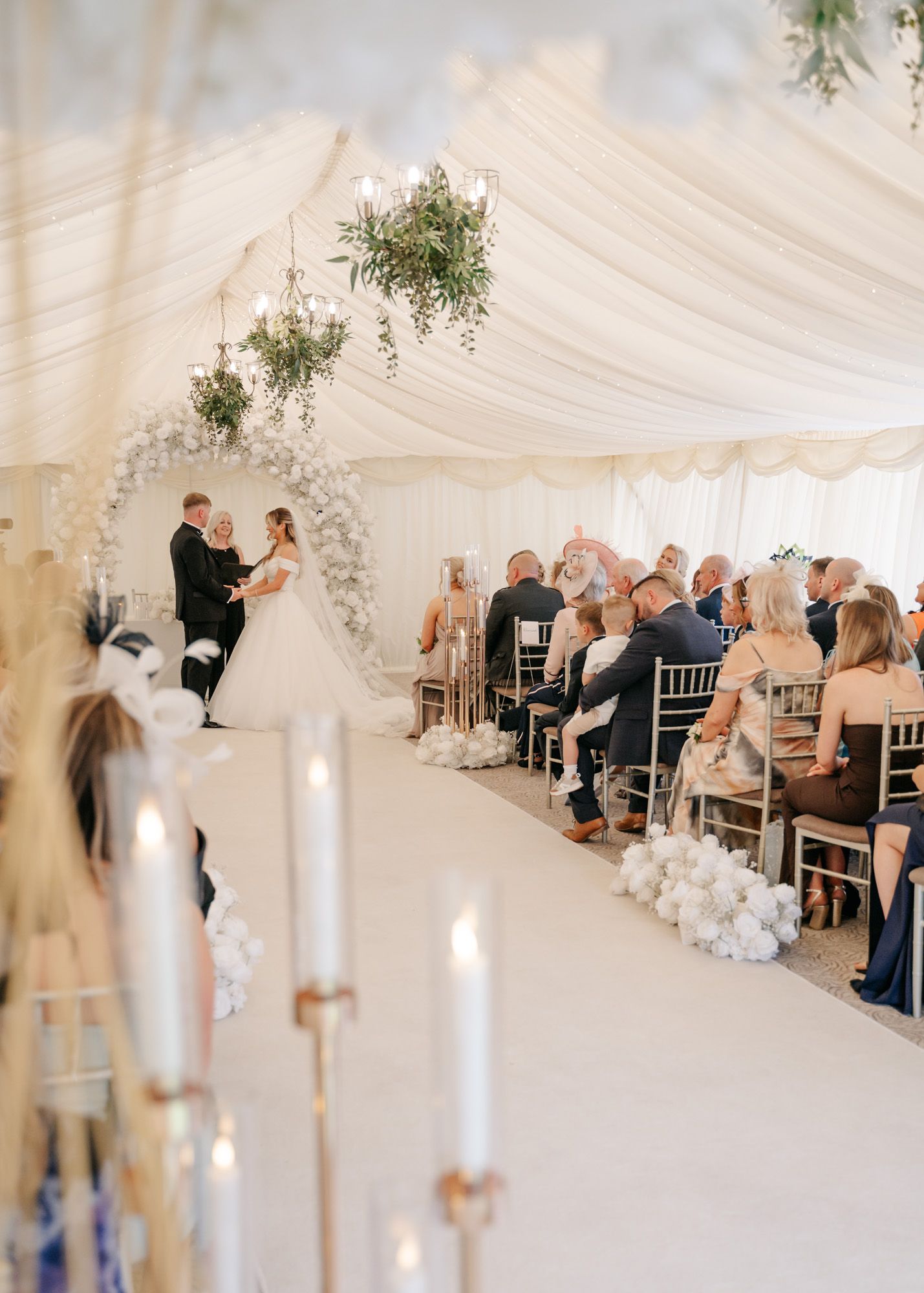 Elegant indoor wedding ceremony under draped white tent with floral arch and candlelit aisle.