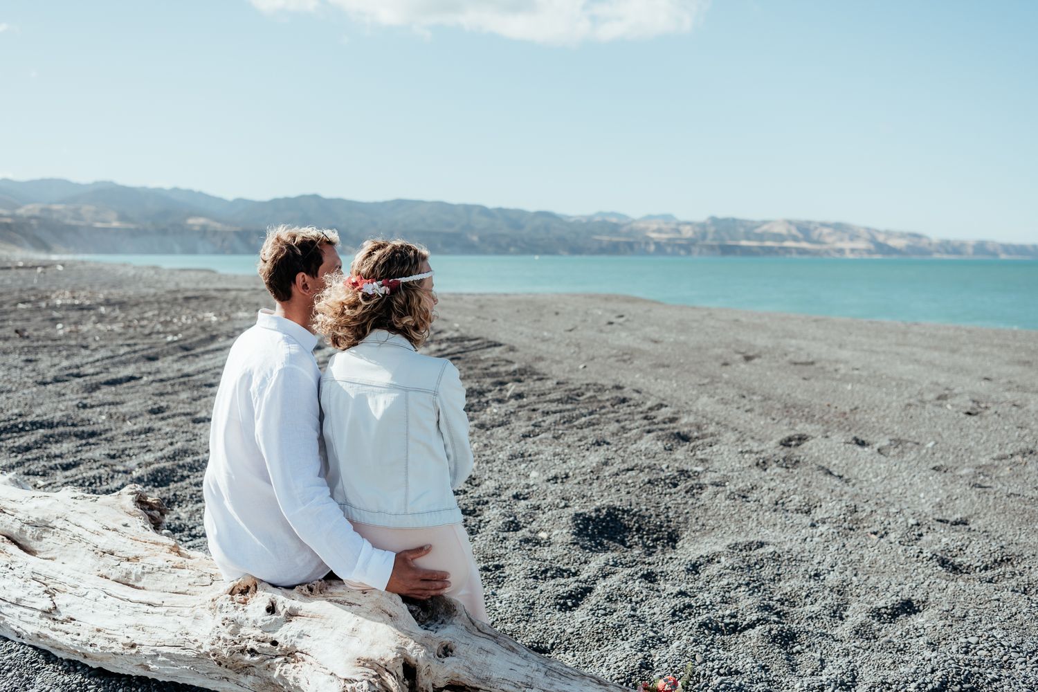An eloping couple sit on a log at Whangaimoana Beach Wairarapa with turquoise water and mountains in the background by August & Vine