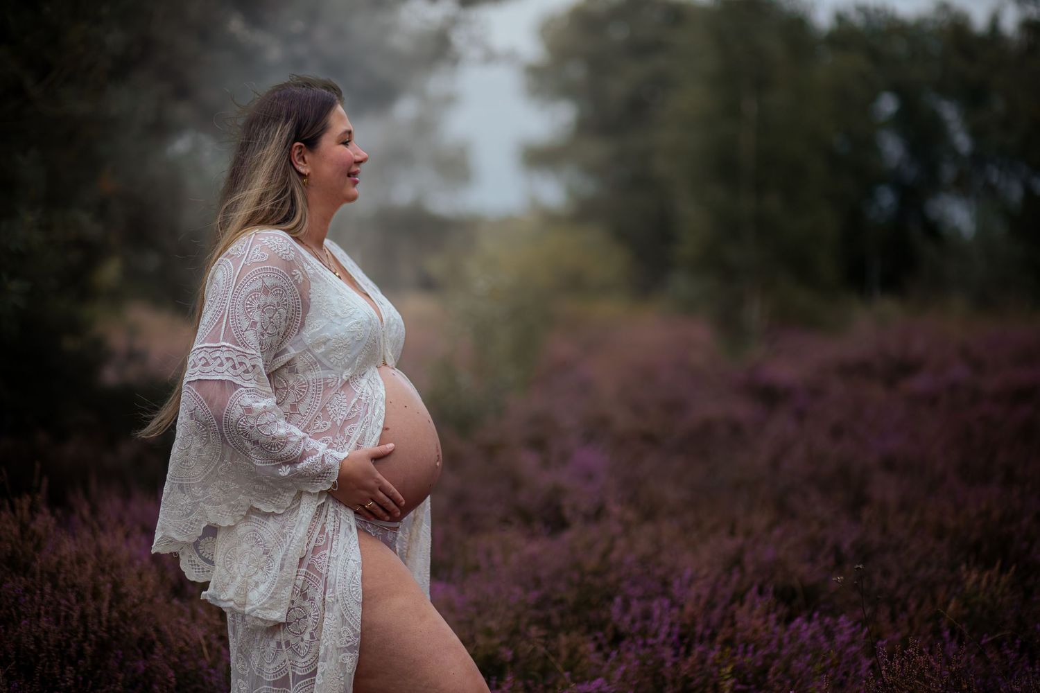 Foto van zwangere dame staand tussen de heide met handen op haar buik.