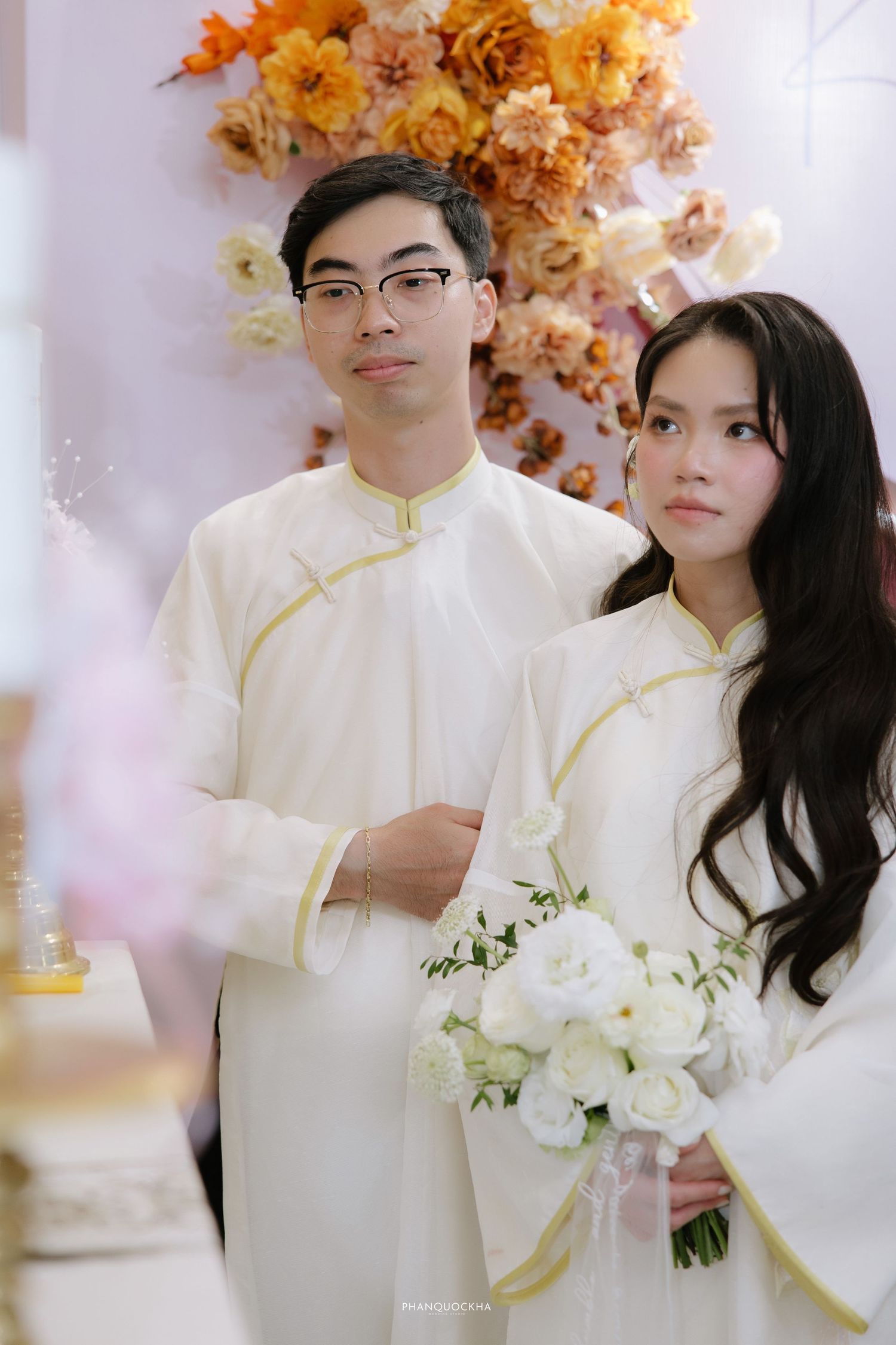 Vietnamese couple in matching white ao dai attire poses with white bouquet before orange floral backdrop.