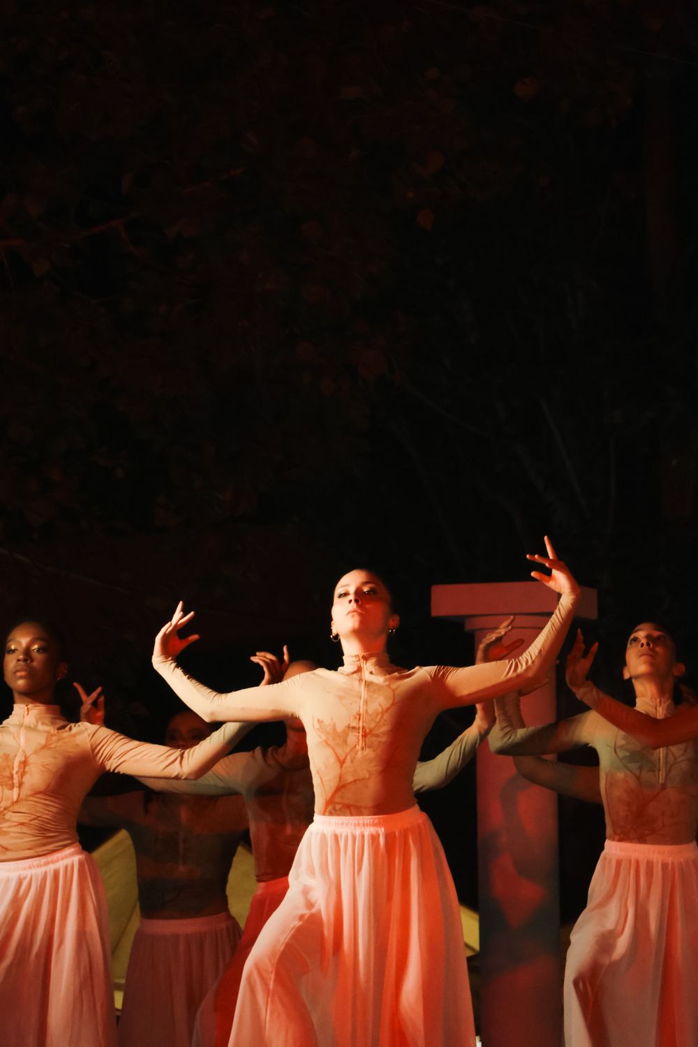 Performers in flowing pink dresses dancing on a dark stage with dramatic lighting.