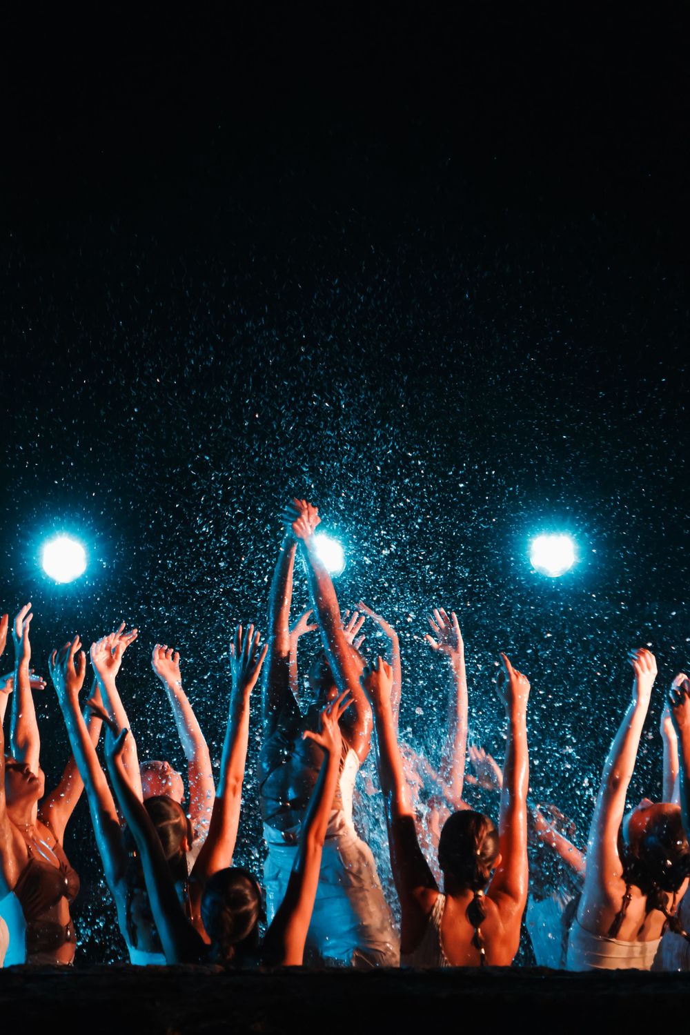 Silhouetted crowd with raised arms at a concert with blue spotlights and glittering atmosphere.